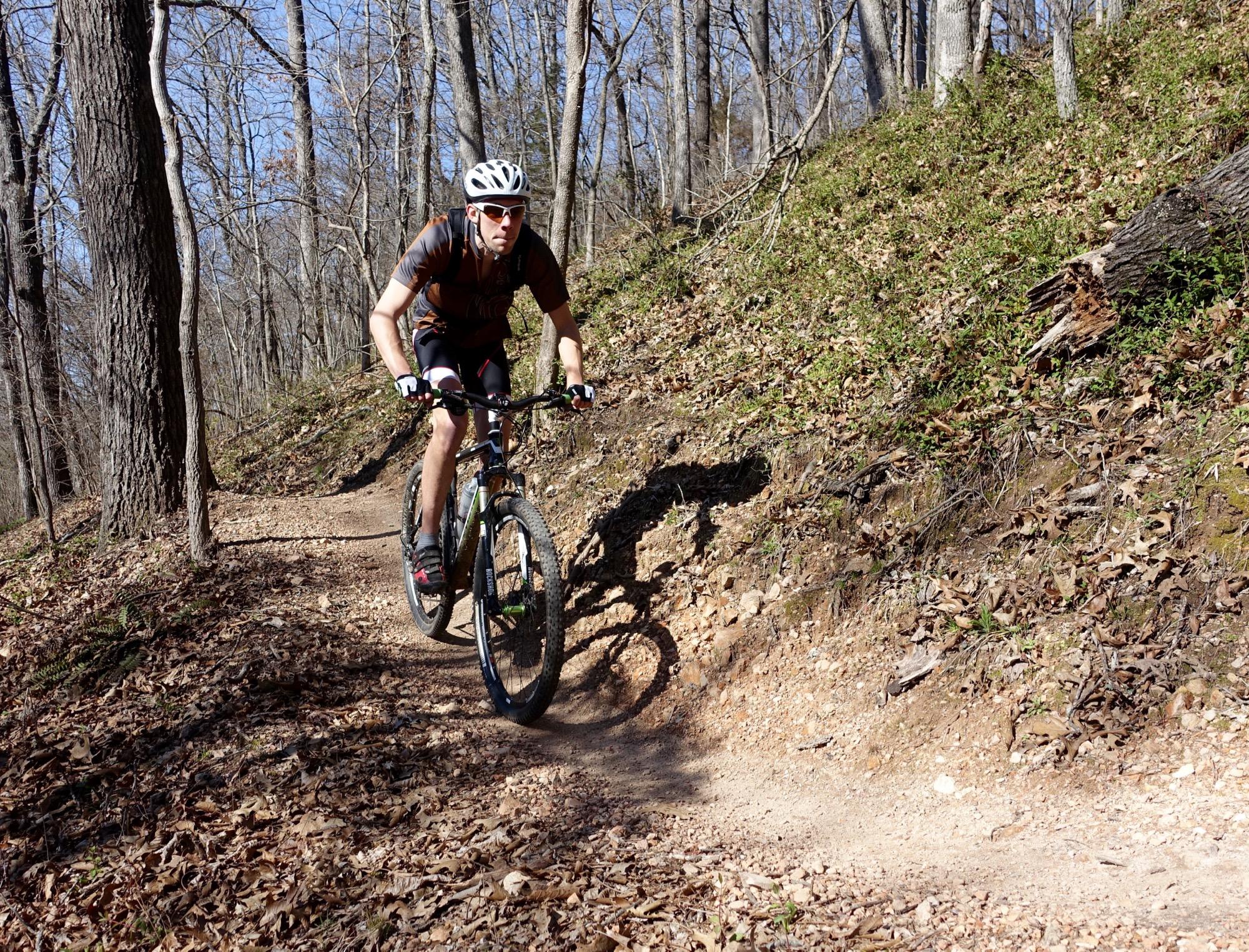 A mountain biker navigating a dirt trail in a wooded area, surrounded by trees and fallen leaves, on a sunny day. The cyclist is wearing a helmet and sunglasses, and appears focused as they ride along the curve of the path. Back 40 mountain bike trail.