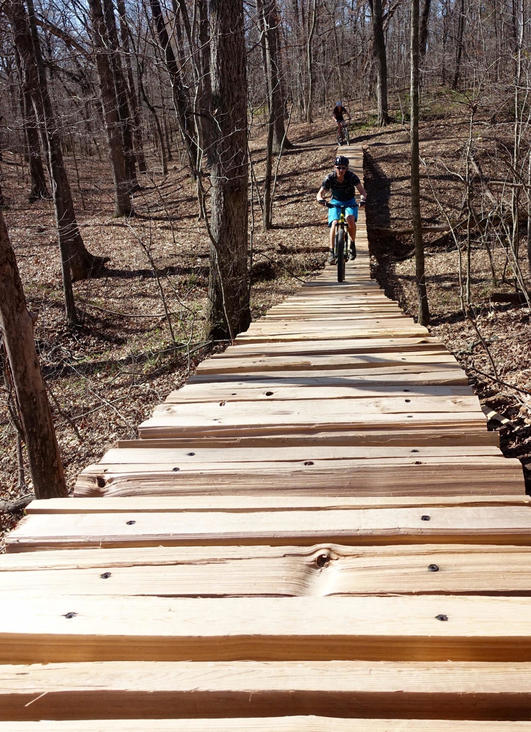 A mountain biker riding along a wooden boardwalk trail in a forested area during daylight. The trail, made of planks, runs downhill with trees and dry leaves visible on either side. Another cyclist is seen in the background, navigating the path. Back 40 mountain bike trail.