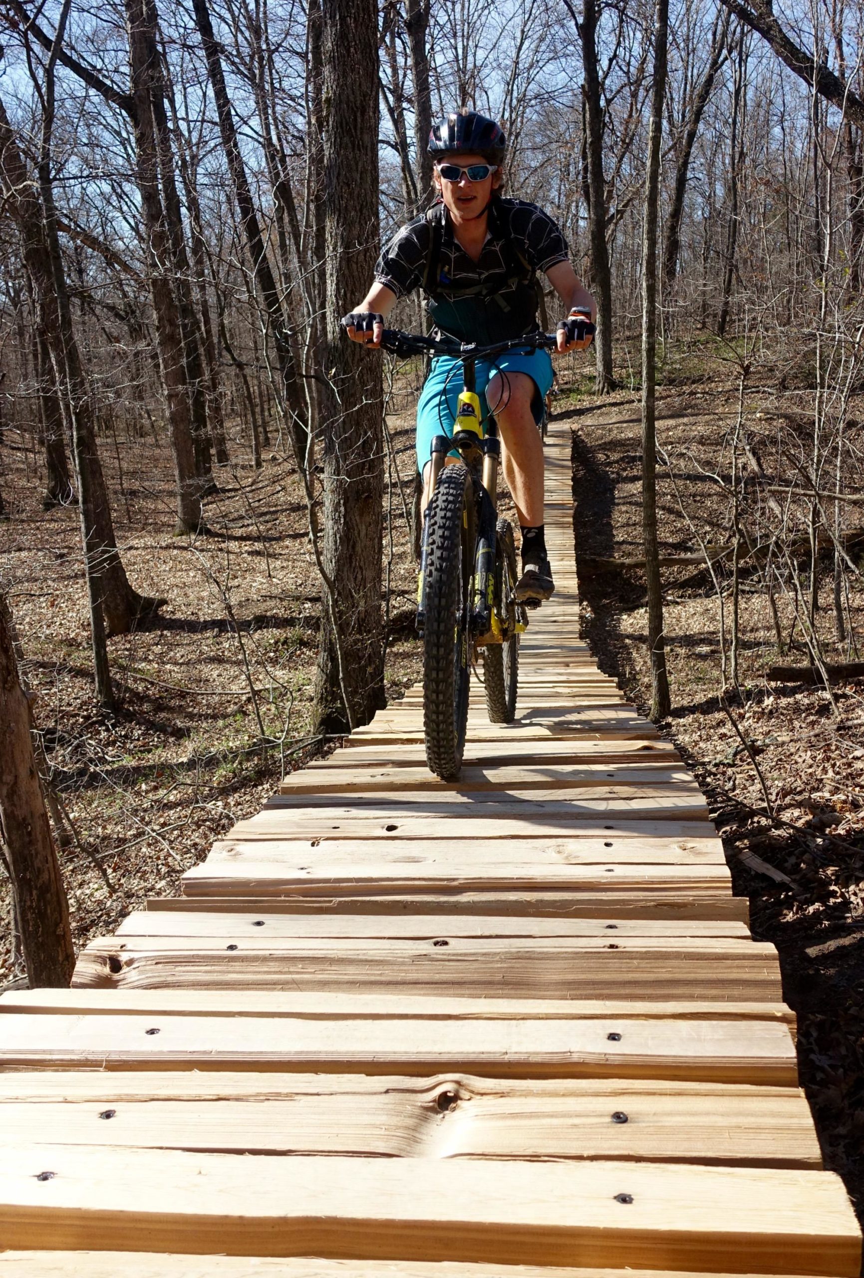 A person riding a mountain bike on a wooden trail through a wooded area. The setting features bare trees in early spring, with the cyclist wearing a helmet, sunglasses, and athletic clothing. Sunlight illuminates the scene, highlighting the wooden planks of the trail. Back 40 mountain bike trail.