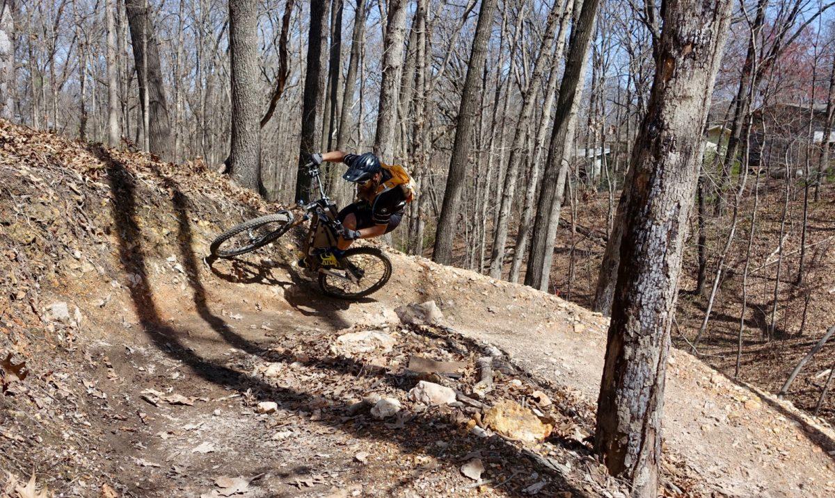 A mountain biker skillfully navigates a winding dirt trail through a wooded area, leaning into a turn as trees rise in the background. The ground is covered with fallen leaves and rocks, with sunlight filtering through the branches. Back 40 mountain bike trail.