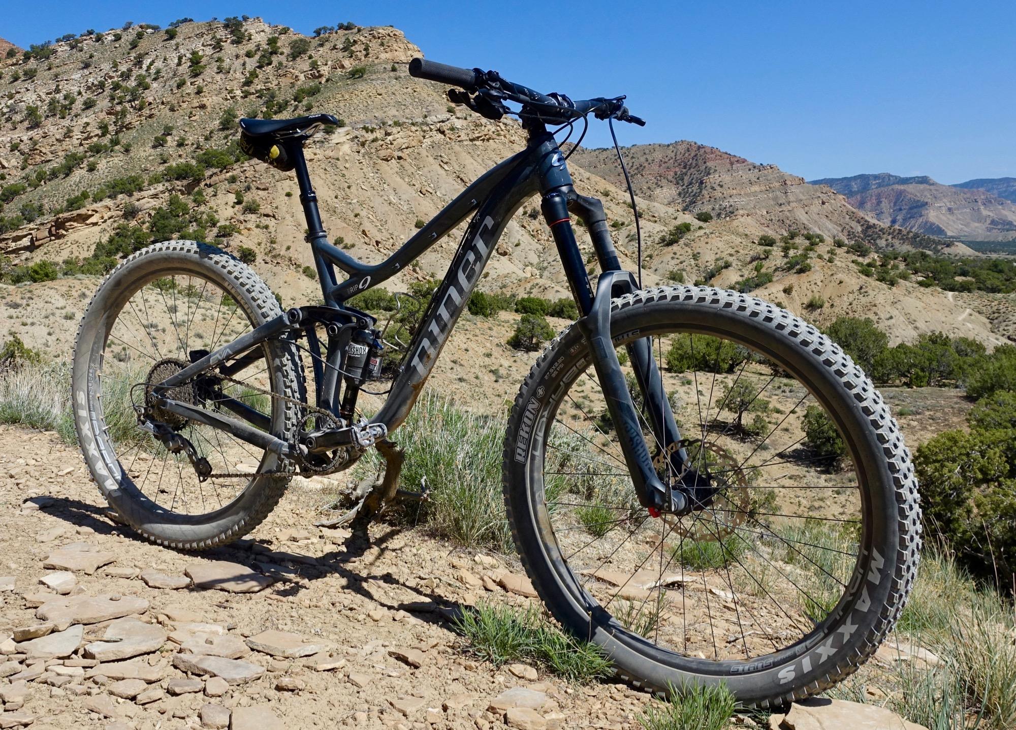 Niner RIP 9: A black mountain bike positioned on a rocky trail, with a backdrop of rolling hills and blue sky. The bike features thick tires and a durable frame, surrounded by sparse vegetation and rugged terrain.
