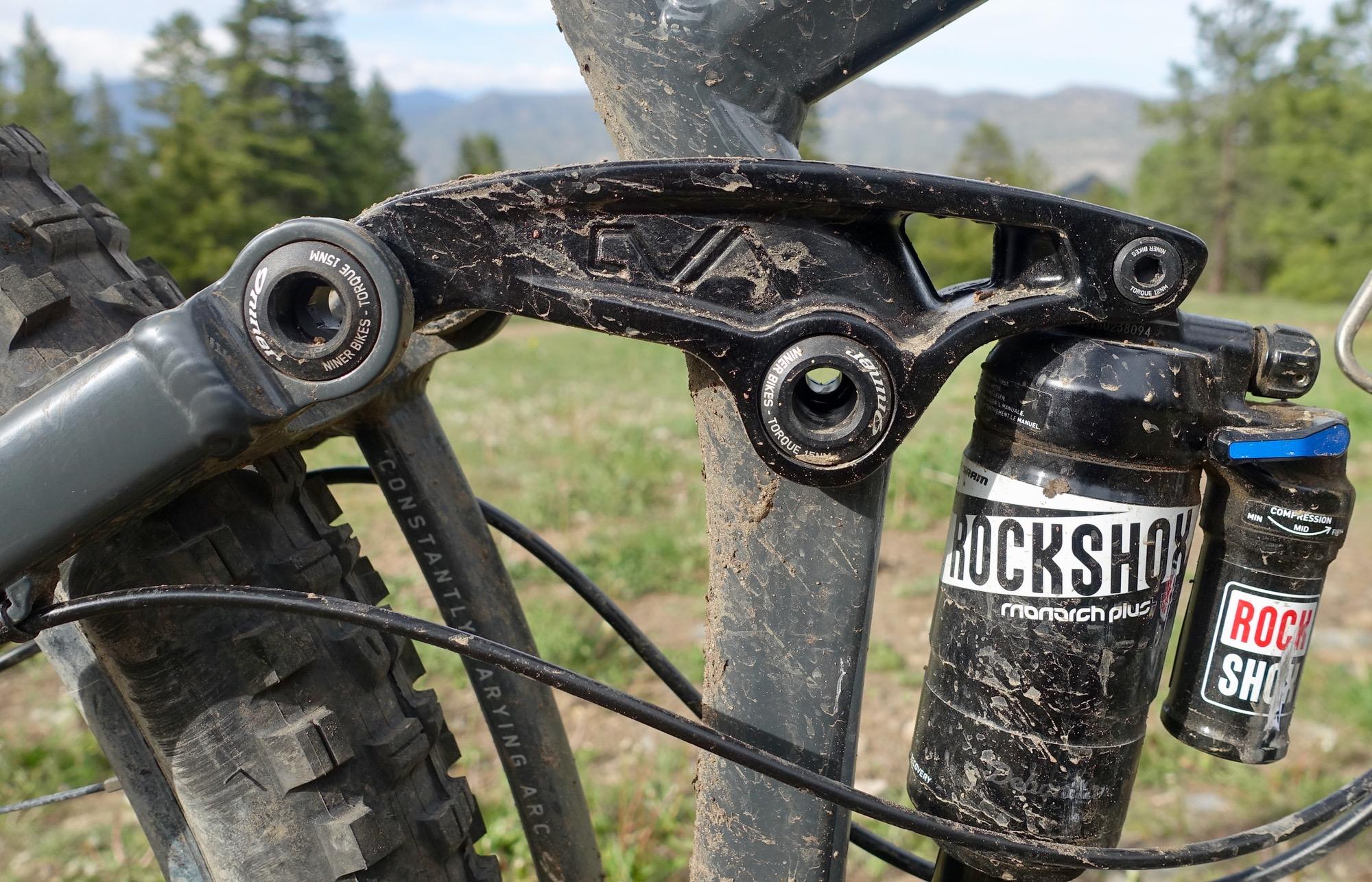 Niner RIP 9: Close-up view of a mountain bike's rear suspension, featuring a black linkage and shock absorber labeled "RockShox Monarch Plus." The bike frame shows some dirt, indicating outdoor use, with a blurred green background suggesting a natural setting.