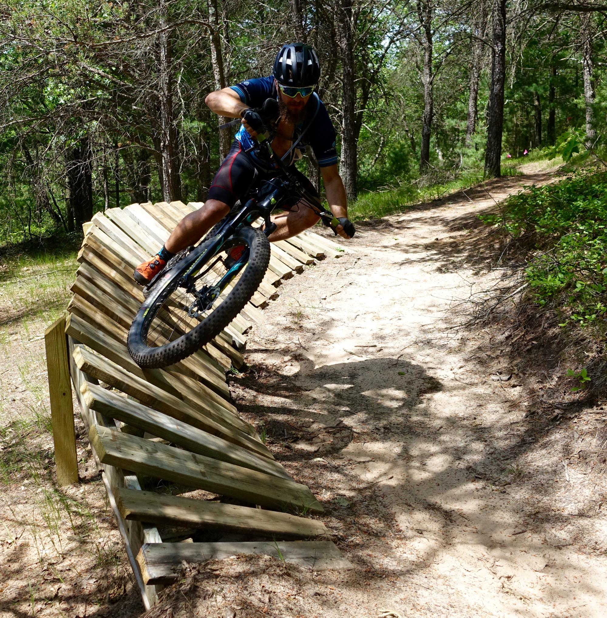 A mountain biker executes a stylish jump on a wooden ramp along a dirt trail, surrounded by trees and greenery. The rider is wearing a helmet and sunglasses, showcasing a dynamic moment in off-road biking. Levis Mounds mountain bike trail.