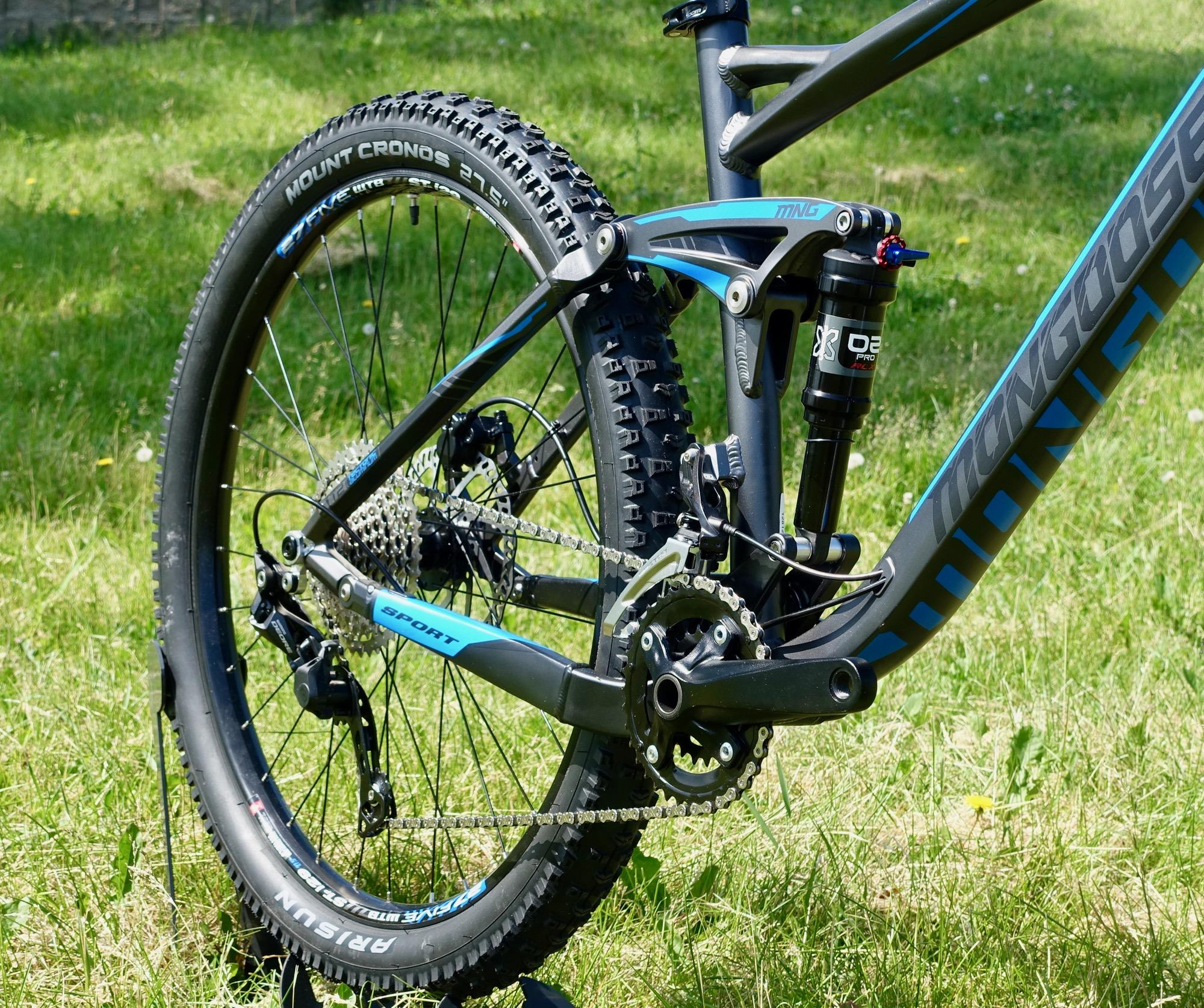 Mongoose Teocali Expert: A close-up view of the rear section of a modern mountain bike, featuring a detailed look at the chain, gears, and suspension system. The bike frame has a sleek design in shades of gray and blue, with visible tire tread on a thick, knobby tire. The background showcases a grassy area, highlighting the outdoor setting.