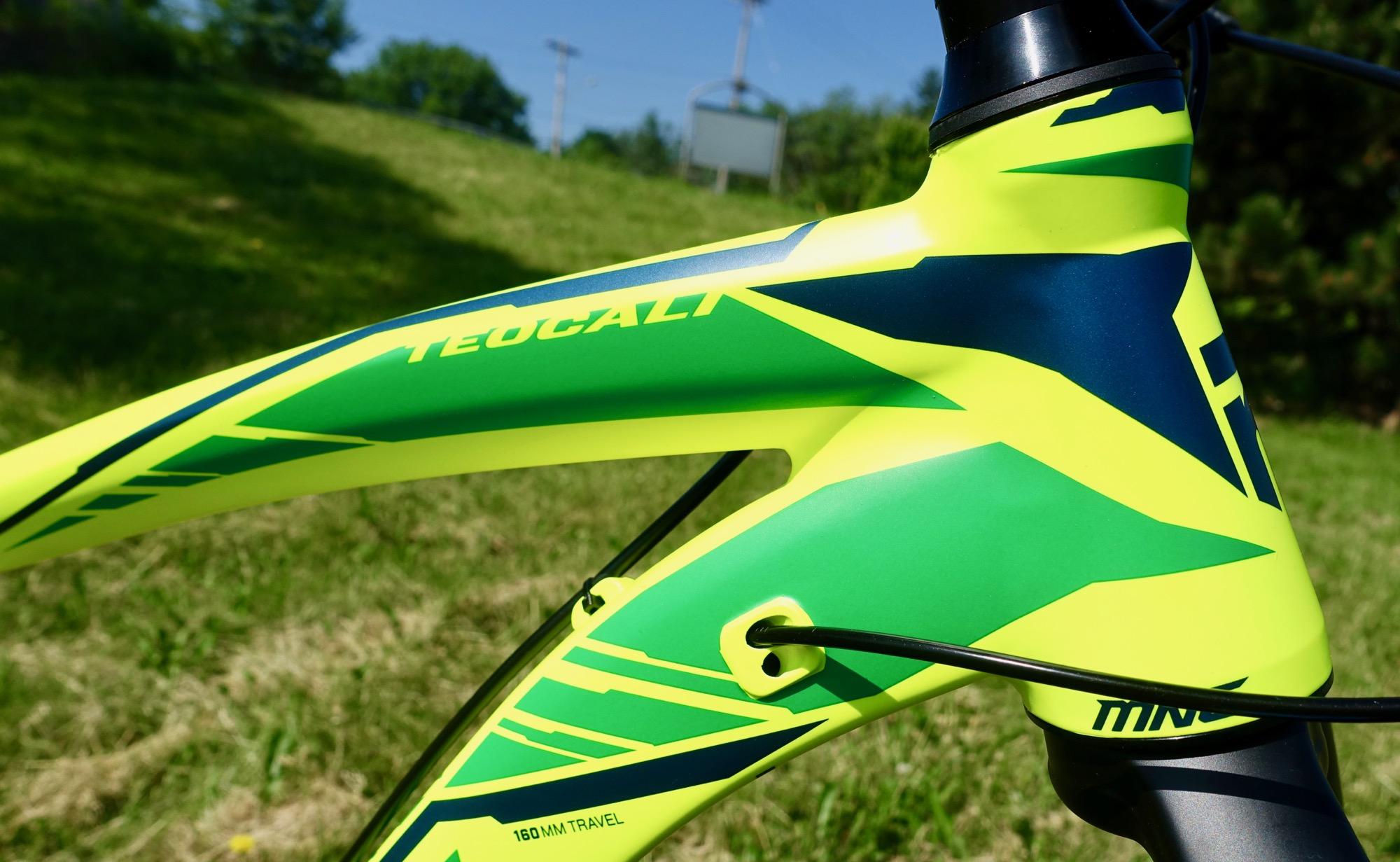 Mongoose Teocali Expert: Close-up view of a brightly colored mountain bike frame featuring a neon green and blue design, with visible cable routing and the model name "Teocali" highlighted on the top tube. The background shows a soft-focus grassy area under clear blue skies.