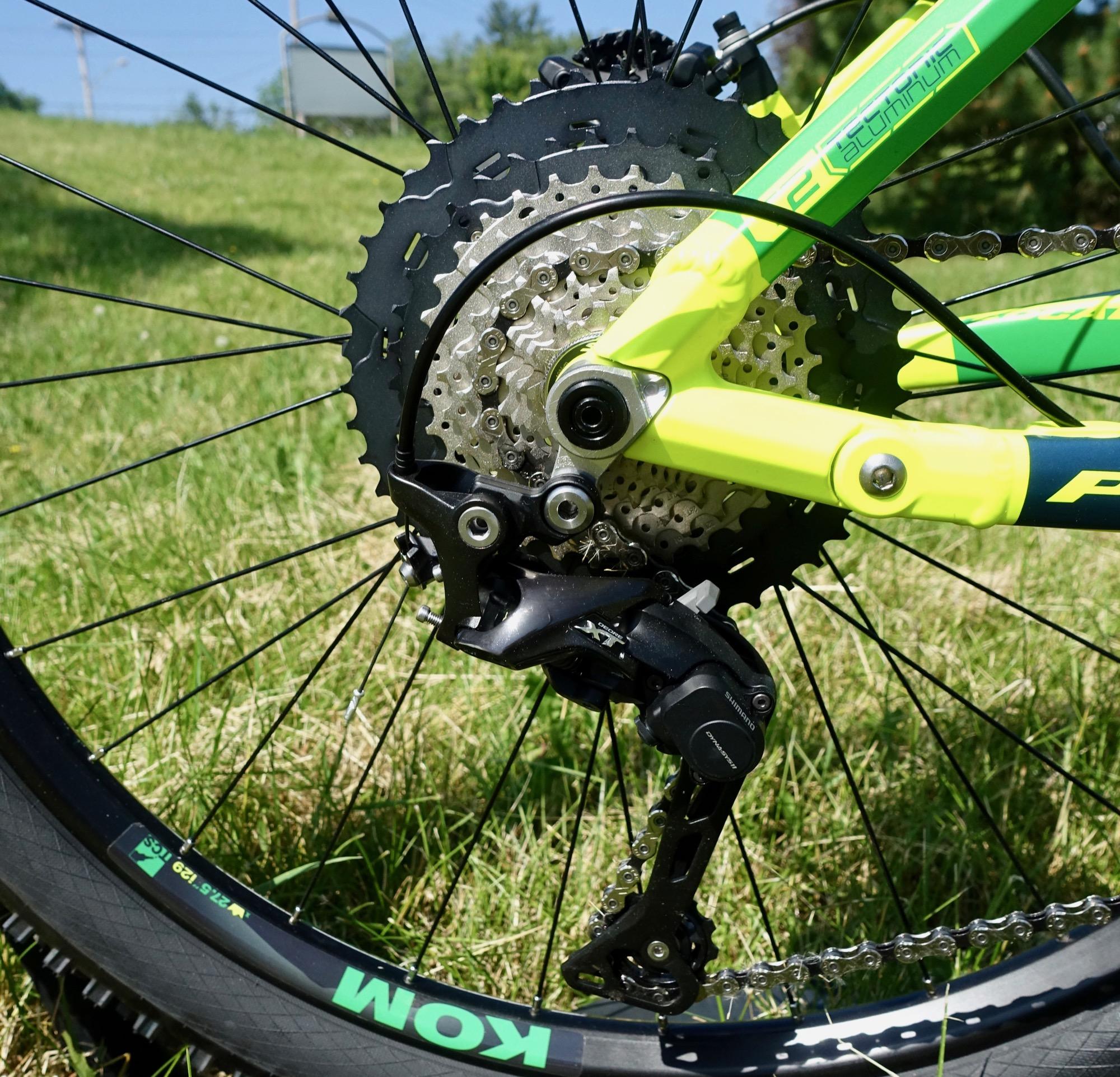 Mongoose Teocali Expert: Close-up of a bicycle's rear drivetrain, showcasing the chain, derailleur, and cassette against a grassy background. The bike features a vibrant green and yellow frame, with the word "KOM" visible on the tire.