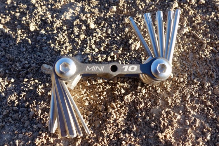 A compact multi-tool with multiple metallic Allen wrench heads, positioned on a textured dirt surface. The tool features a central black handle with "MINI" and "10" printed on it, surrounded by the unfolded tool components.