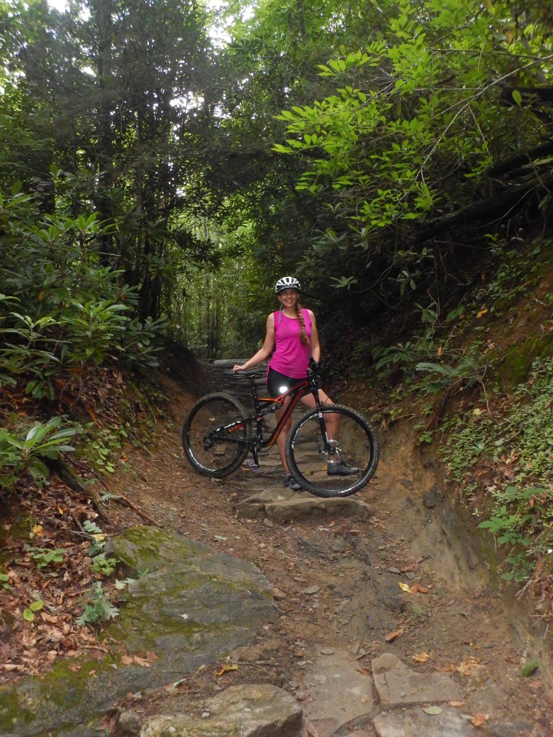 A person wearing a pink tank top and a helmet stands beside a mountain bike on a rocky trail surrounded by lush greenery. The path is narrow, flanked by dense foliage, creating a serene, natural setting. Black Mountain mountain bike trail.