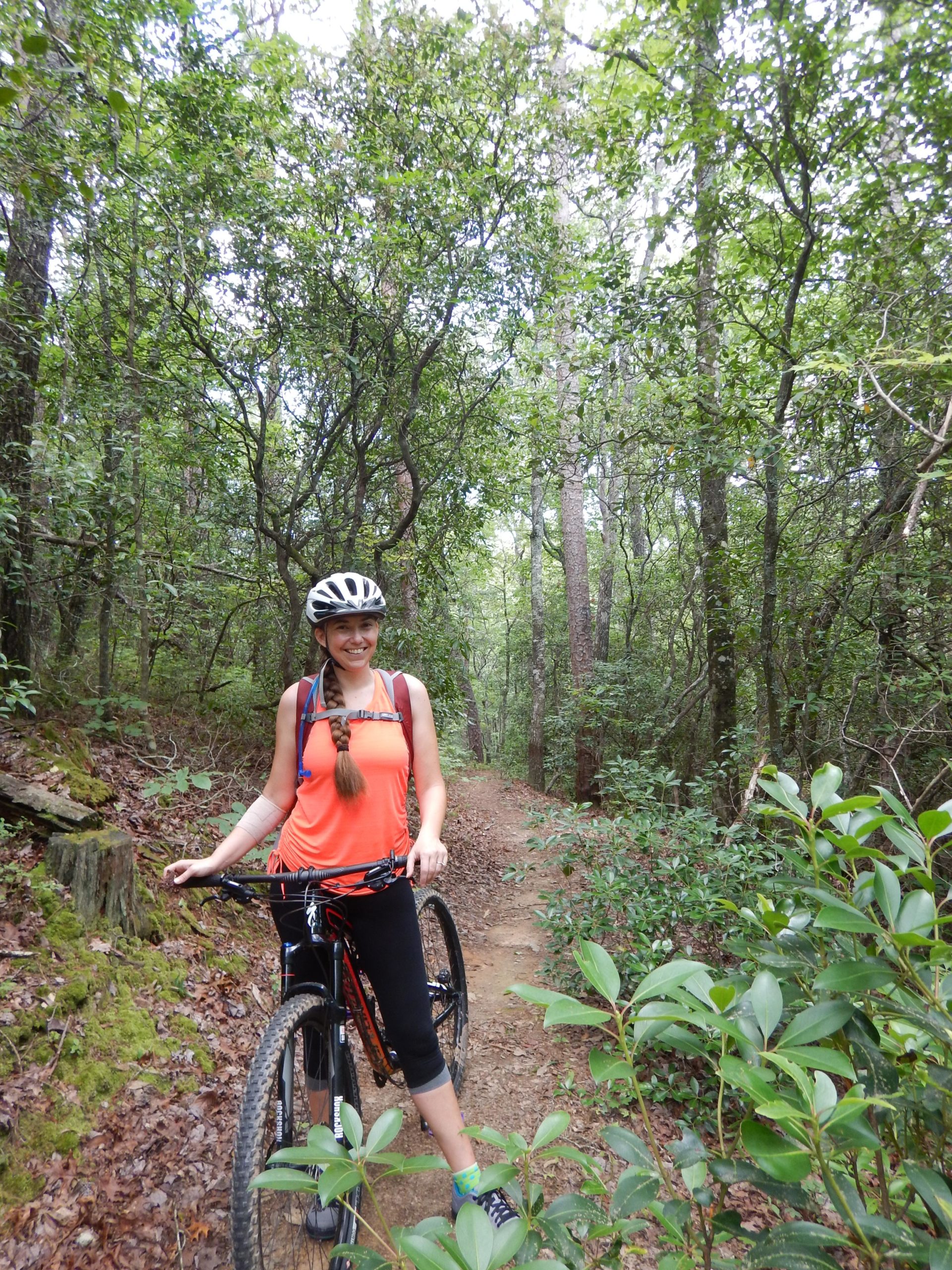 A young woman stands on a dirt biking trail in a lush green forest, holding her mountain bike. She is wearing a bright orange tank top, black leggings, and a bicycle helmet. The surroundings include trees, shrubs, and a winding path, creating an outdoor adventure setting. Stonewall Falls Loop mountain bike trail.