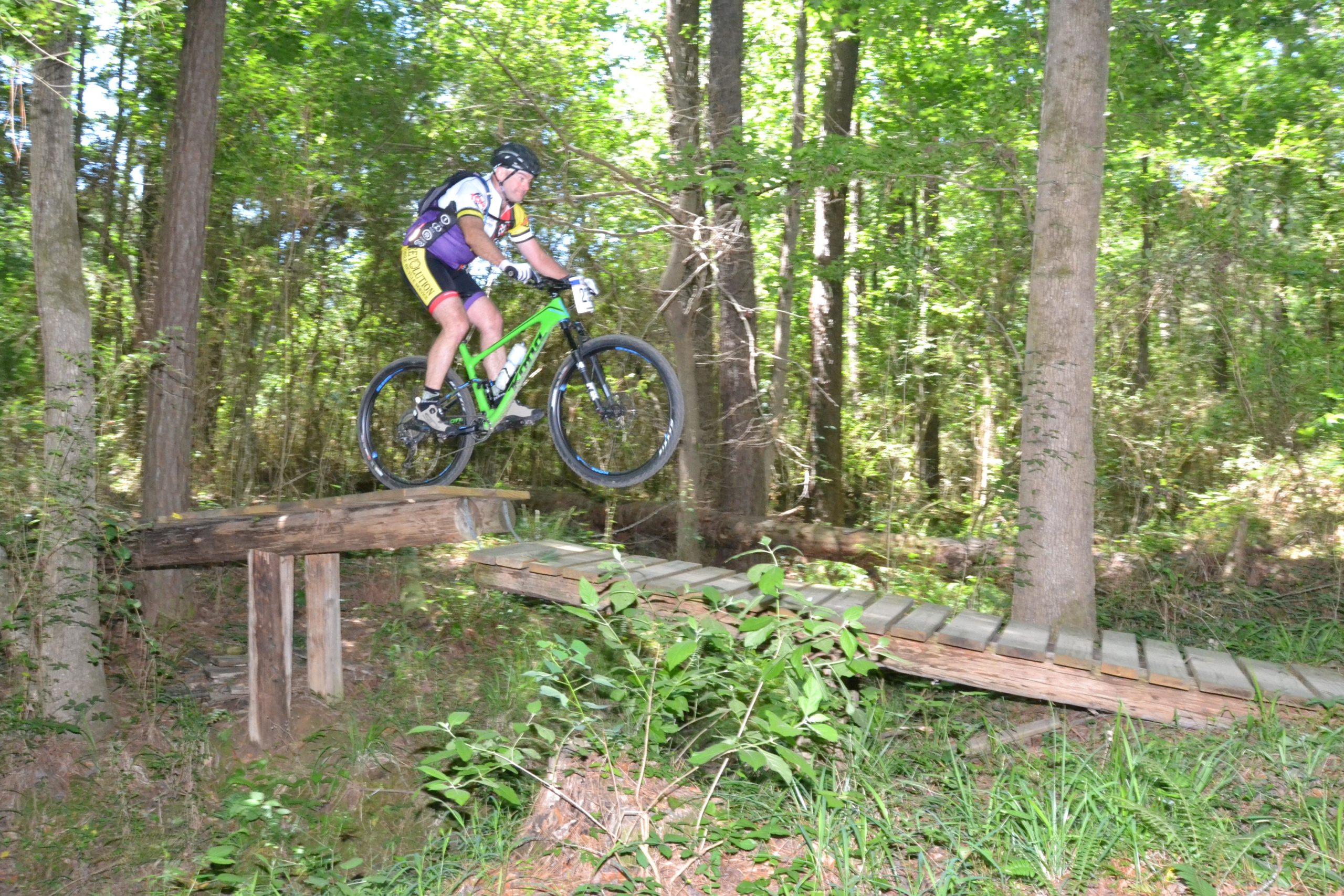 A mountain biker in athletic gear jumps over a wooden bridge in a forested area, surrounded by tall trees and greenery. The bike is angled mid-air, showcasing the rider's skill as they navigate a dirt trail. Mt. Zion Bike Trails mountain bike trail.