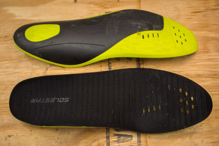 Black and yellow shoe insoles, one side featuring a textured black surface with the word "SoleStar" visible, and the other side showing a shiny yellow and black design with ventilation holes. The insoles are placed on a wooden surface.