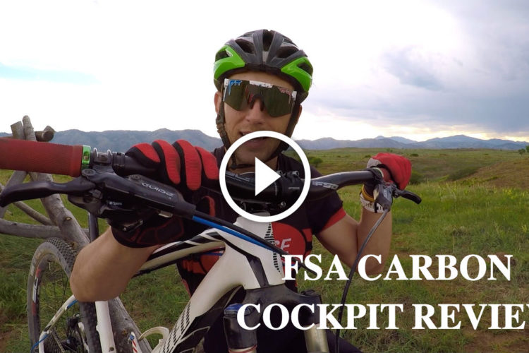 A person wearing a helmet and sunglasses is seated on a mountain bike, showcasing the handlebars and controls. The background features a scenic view of grassy hills and mountains, with text overlay indicating a video review for "FSA Carbon Cockpit."