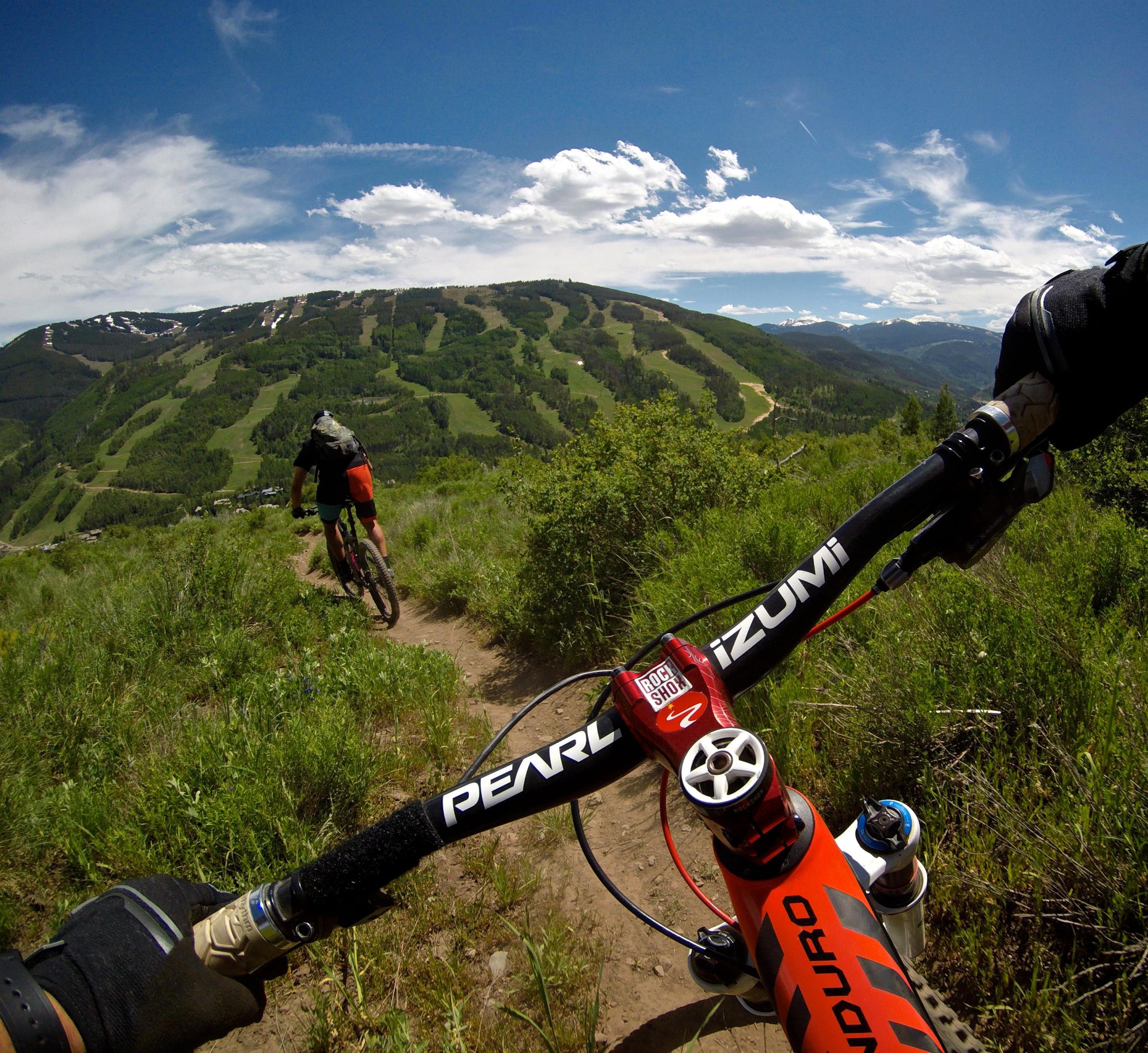 Mountain biker riding downhill on a dirt trail, with a scenic view of a lush green hillside and cloudy blue sky in the background. The perspective shows the handlebars of the bike in the foreground. Son Of Middle Creek mountain bike trail.
