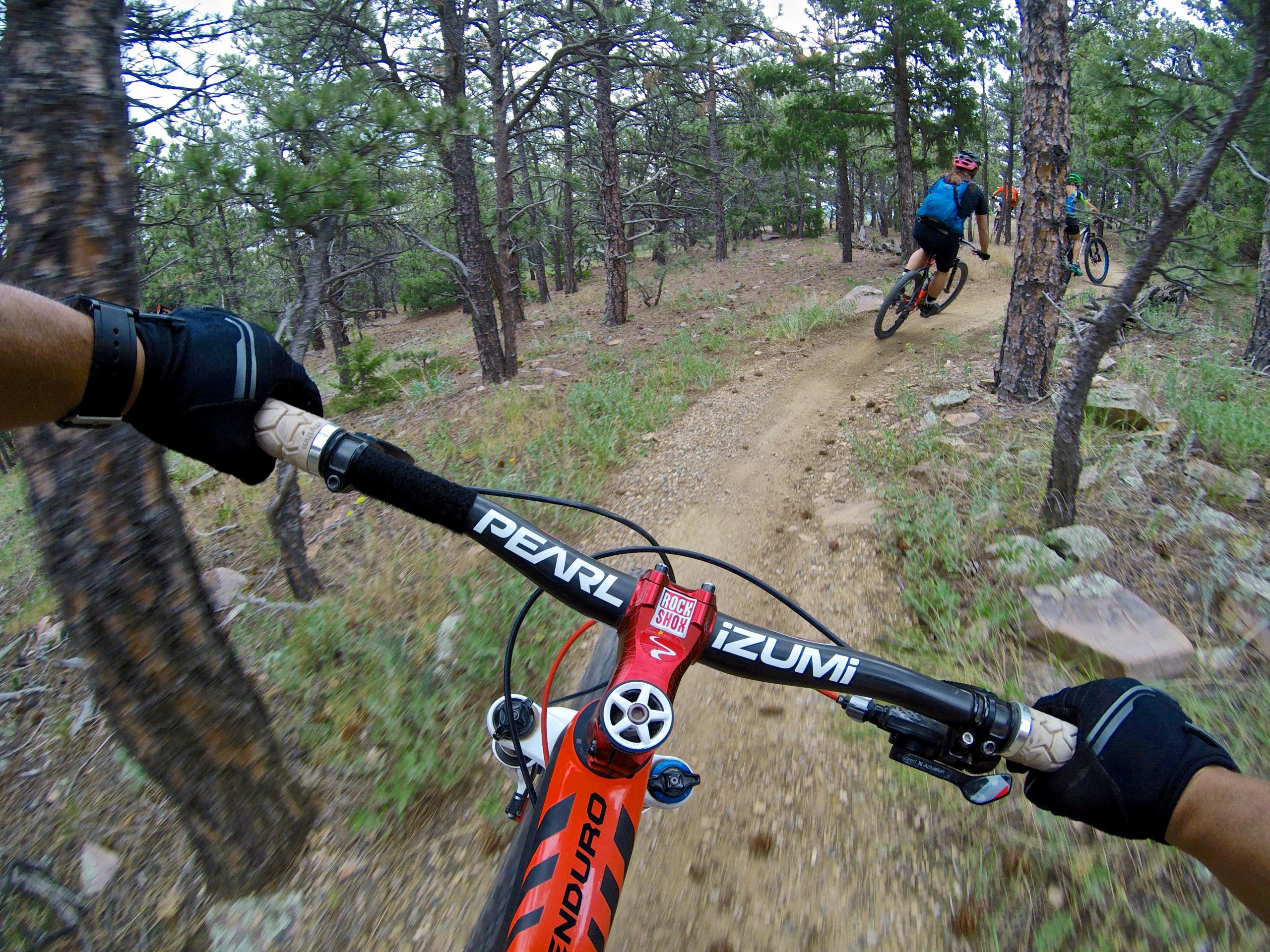 A cyclist's perspective while riding a mountain bike on a dirt trail surrounded by trees. The image shows the handlebars of the bike in the foreground, with a gloved hand gripping the handlebars. In the background, other cyclists are visible riding along the winding trail. The scene captures a sense of adventure and the natural beauty of the forested landscape. Heil Valley Ranch mountain bike trail.