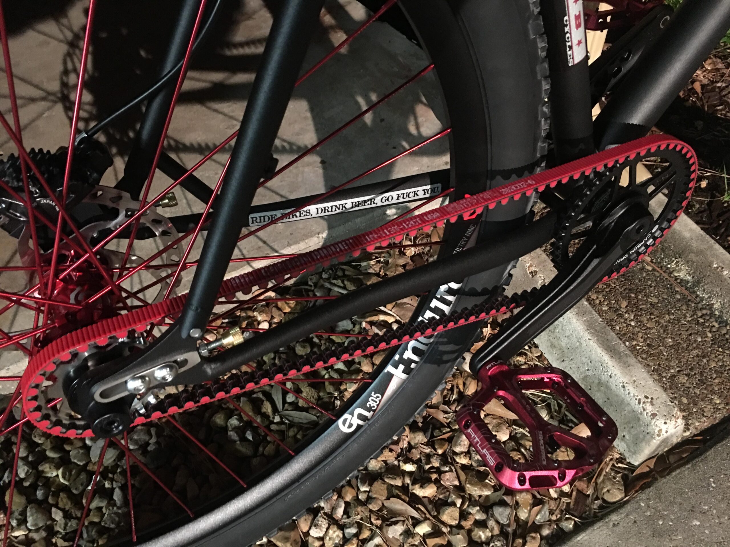 Reeb REEBdikeylous: Close-up of a bicycle's drivetrain, featuring a red belt drive system, black chainring, and a pink pedal. The background shows textured gravel and a concrete curb, with a mixture of shadows indicating nighttime conditions.