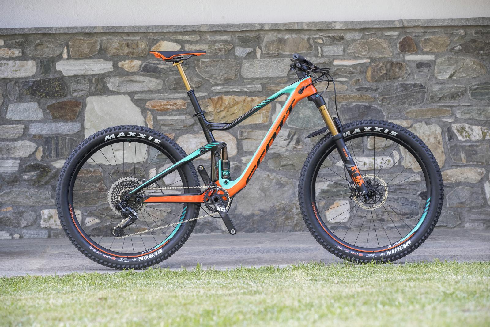 Scott Genius: A brightly colored mountain bike with a black, orange, and turquoise frame is displayed against a stone wall. The bike features large Maxxis tires, a prominent suspension fork, and a sleek design, emphasizing its suitability for off-road riding. The setting includes a patch of green grass beneath the bike.