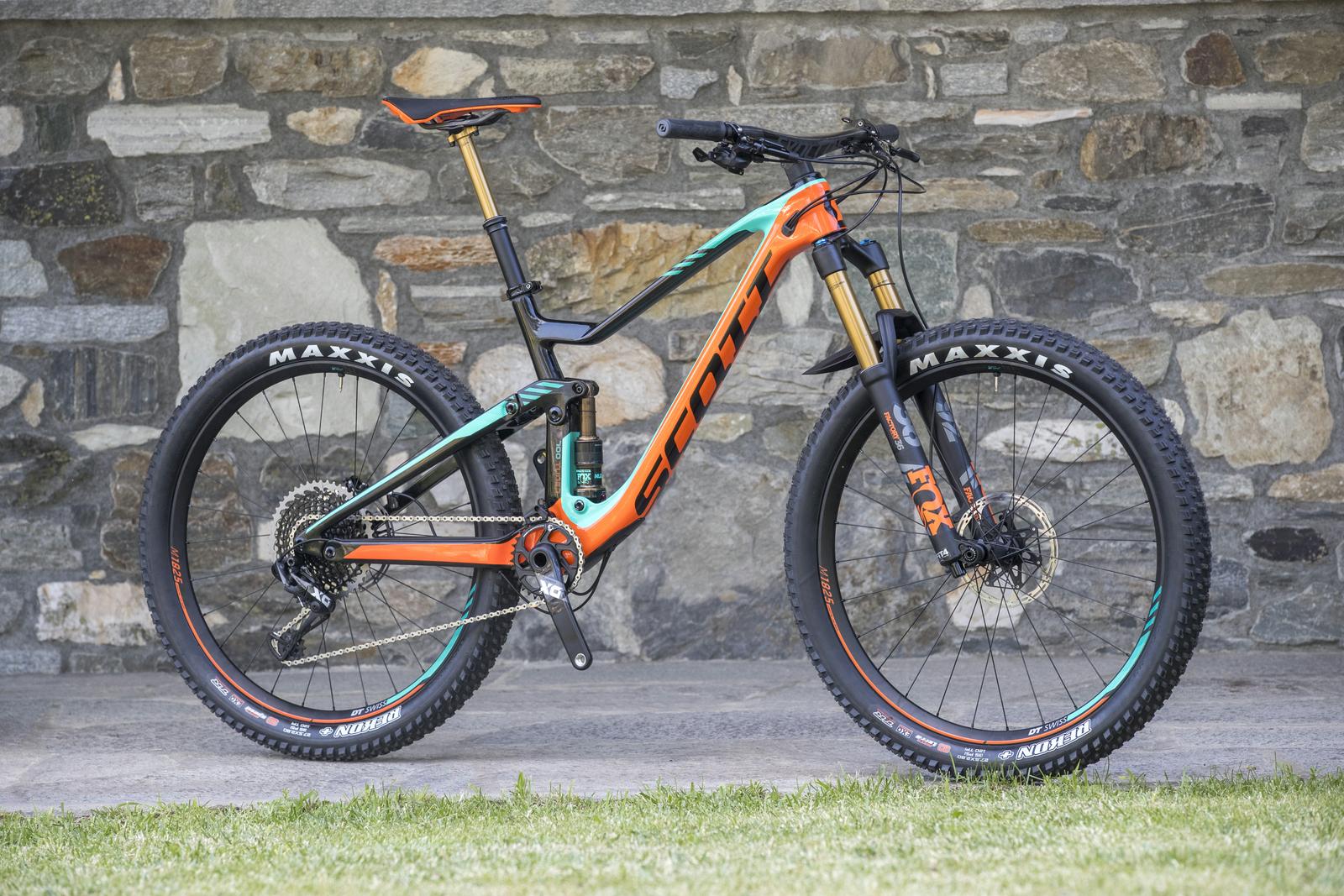 Scott Genius: A modern mountain bike with a vibrant orange and teal color scheme, featuring a lightweight carbon frame, suspension fork, and wide tires mounted on Maxxis rims. The bike is positioned on a stone-covered surface with grass nearby, showcasing its design and components.