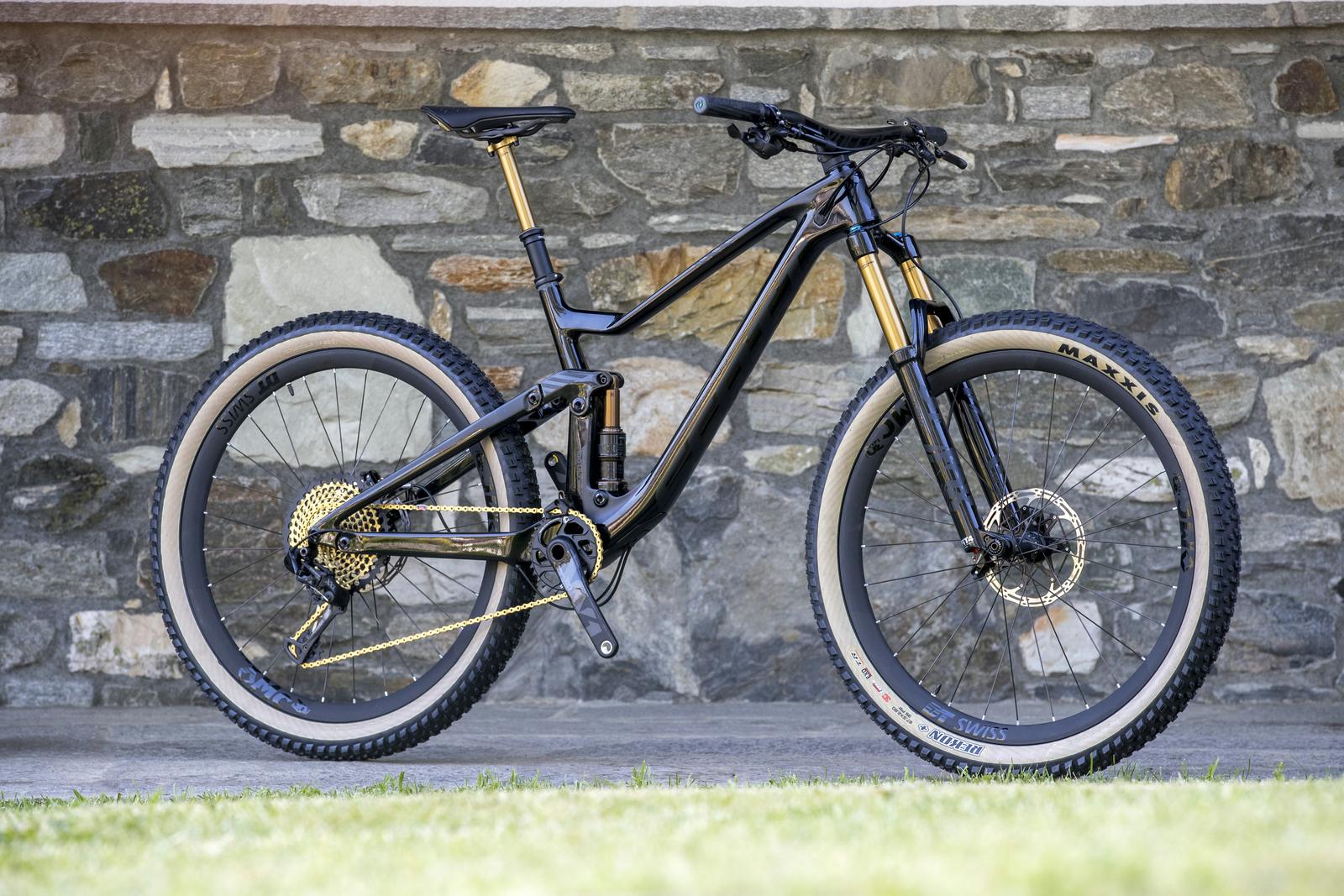 Scott Genius: A black and gold mountain bike stands upright against a stone wall. The bike features a sleek design with a prominent suspension system, detailed gears, and chunky tires branded with the name "Maxxis." The setting includes a patch of green grass beneath the bike, highlighting its rugged style and outdoor capability.
