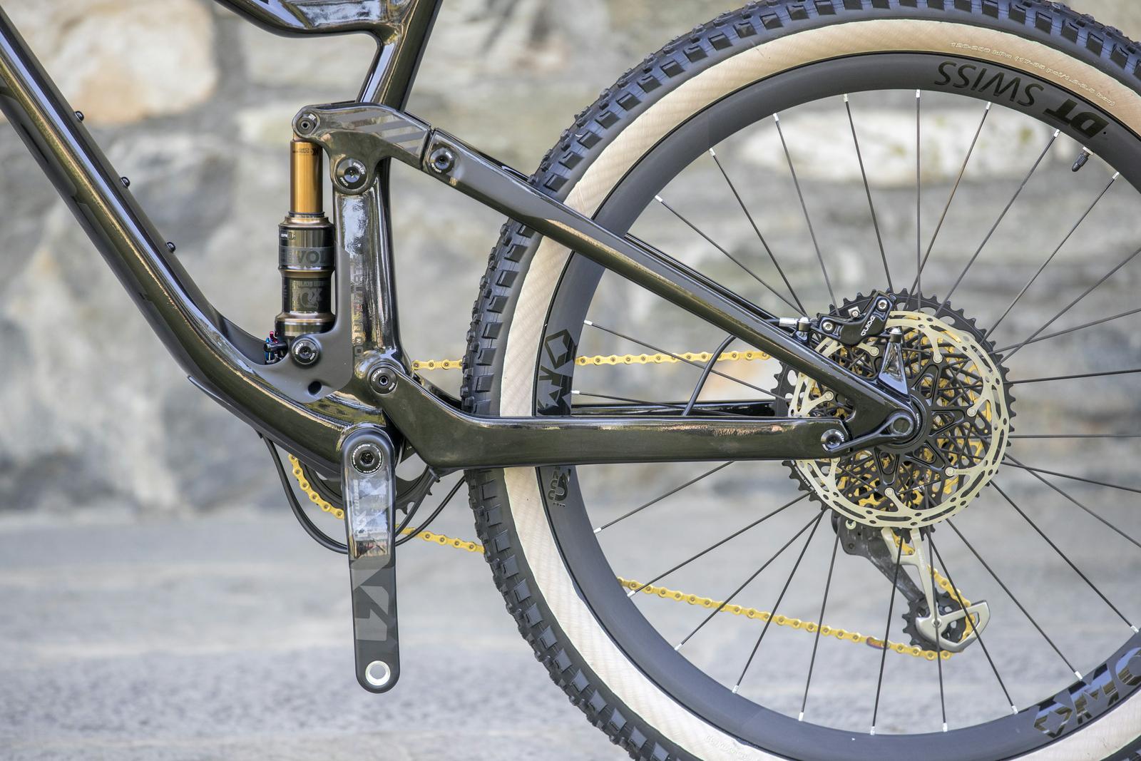 Scott Genius: A close-up view of a mountain bike's rear frame, showcasing the suspension system, gears, and a black tire with a gold chain. The background features a textured stone surface.