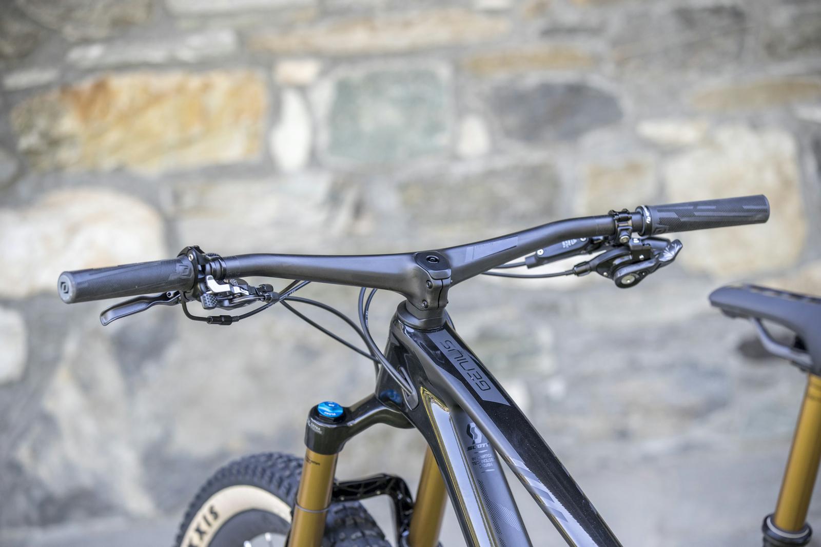 Scott Genius: Close-up view of a mountain bike's handlebars and stem, featuring black grips, brake levers, and a sleek frame, set against a stone wall background.