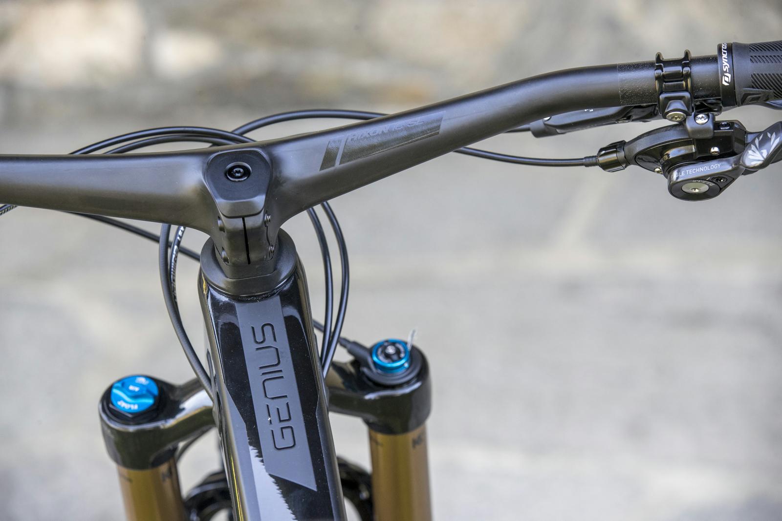Scott Genius: Close-up view of a mountain bike's handlebars, featuring a sleek, black design with the word "GENIUS" prominently displayed. The image showcases the stem, headset, and integrated controls, with cables neatly routed. A blue adjustment knob can be seen on the forks, suggesting advanced suspension features. The background is soft-focused stone pavement, adding context to the outdoor setting.