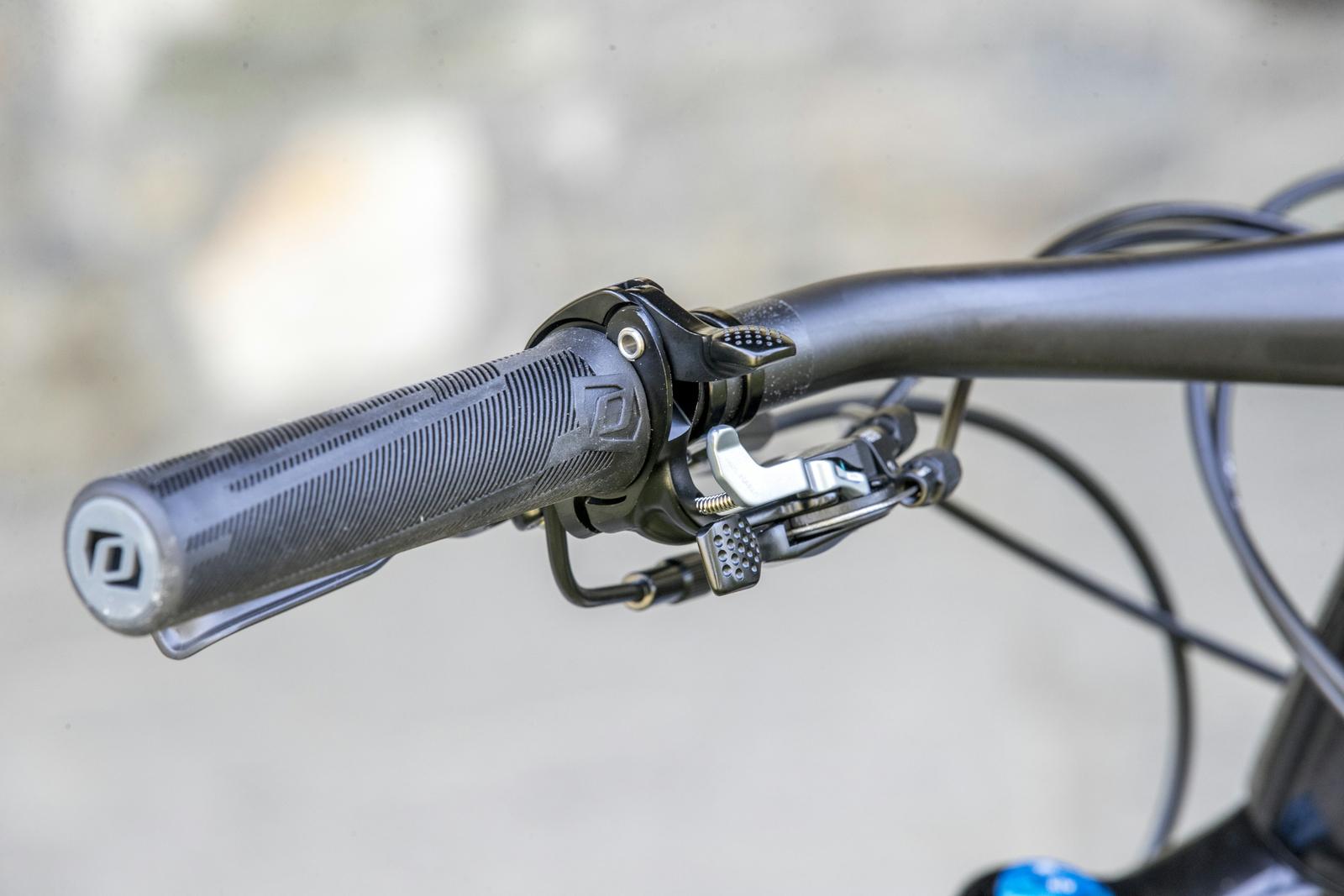 Scott Genius: Close-up view of a mountain bike handlebar featuring a textured grip, gear shifter, and brake lever, set against a blurred background.