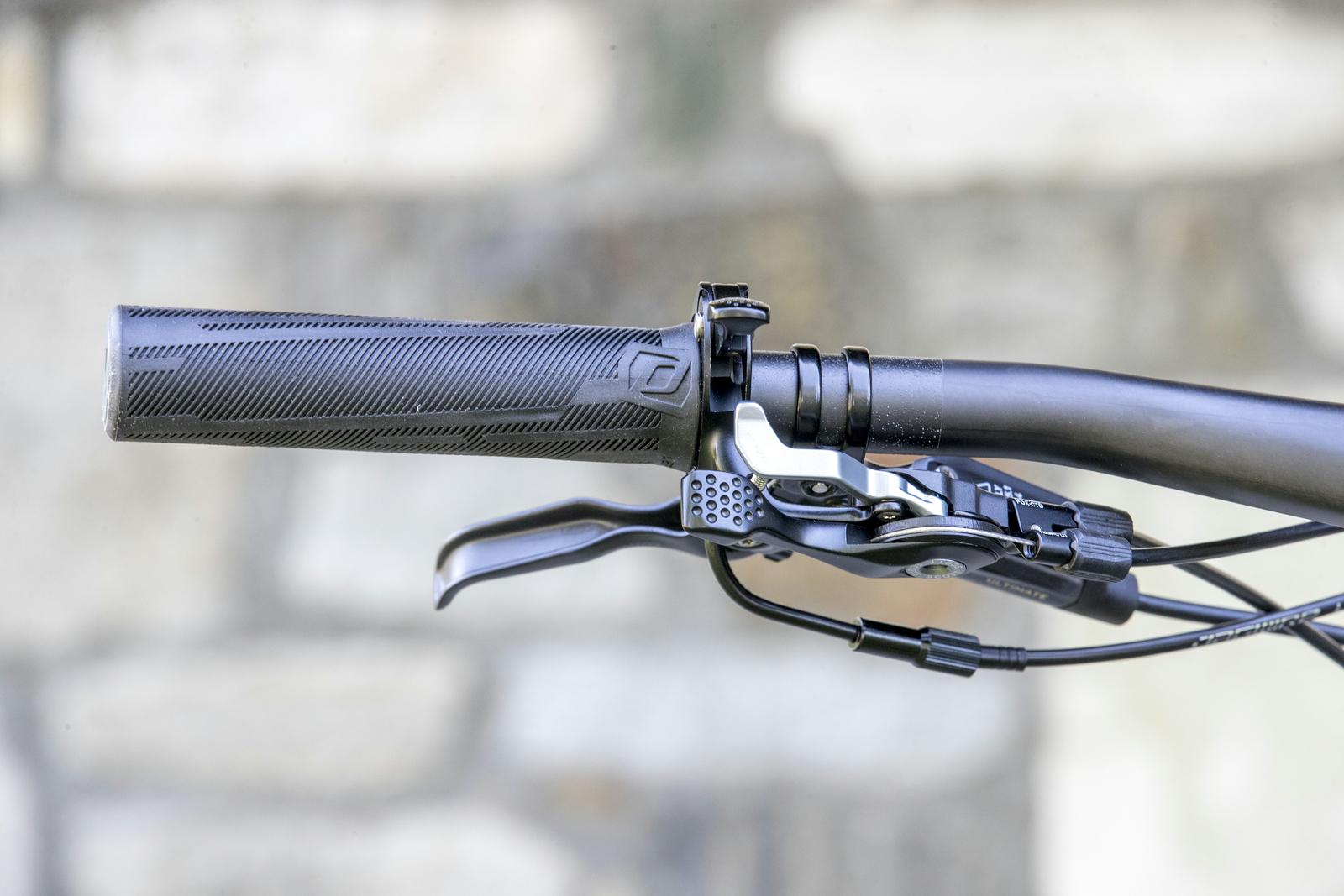 Scott Genius: Close-up view of a bicycle handlebar, featuring a textured black grip, a brake lever, and a gear shift lever, set against a blurred stone background.