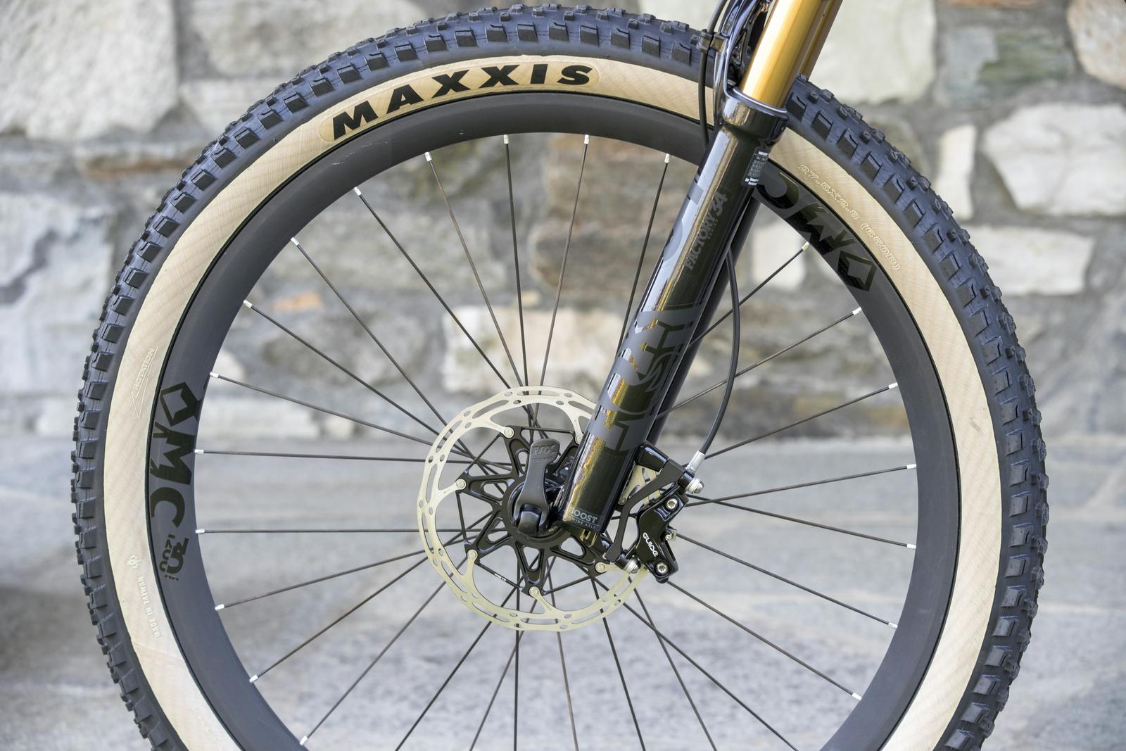 Scott Genius: Close-up view of the front wheel of a mountain bike, featuring a prominent Maxxis tire with a distinctive tread pattern. The wheel includes a gold and black fork and a disc brake system, set against a textured stone background.
