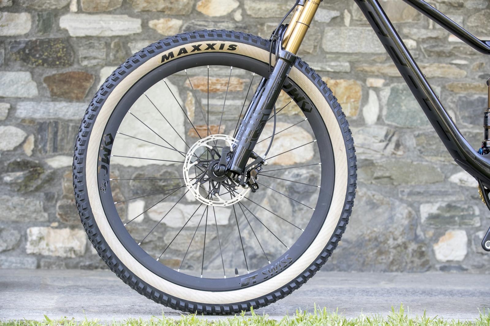 Scott Genius: Close-up image of the front wheel of a mountain bike, featuring a black and gold suspension fork, black spokes, and a distinctive tire with a light tan sidewall and a tread pattern designed for traction. The background consists of a textured stone wall, providing a natural outdoor setting.