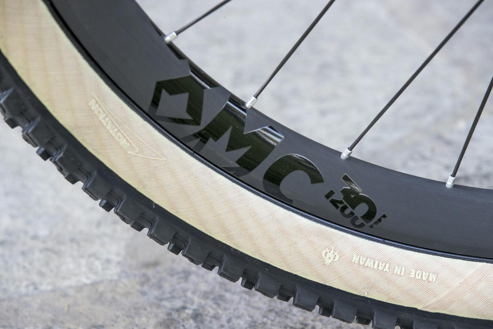 Scott Genius: Close-up image of a mountain bike tire and wheel rim, featuring a distinct black rim with raised lettering and a textured tan sidewall. The tire shows a rugged tread pattern suitable for off-road terrain. The background is a blurred concrete surface.