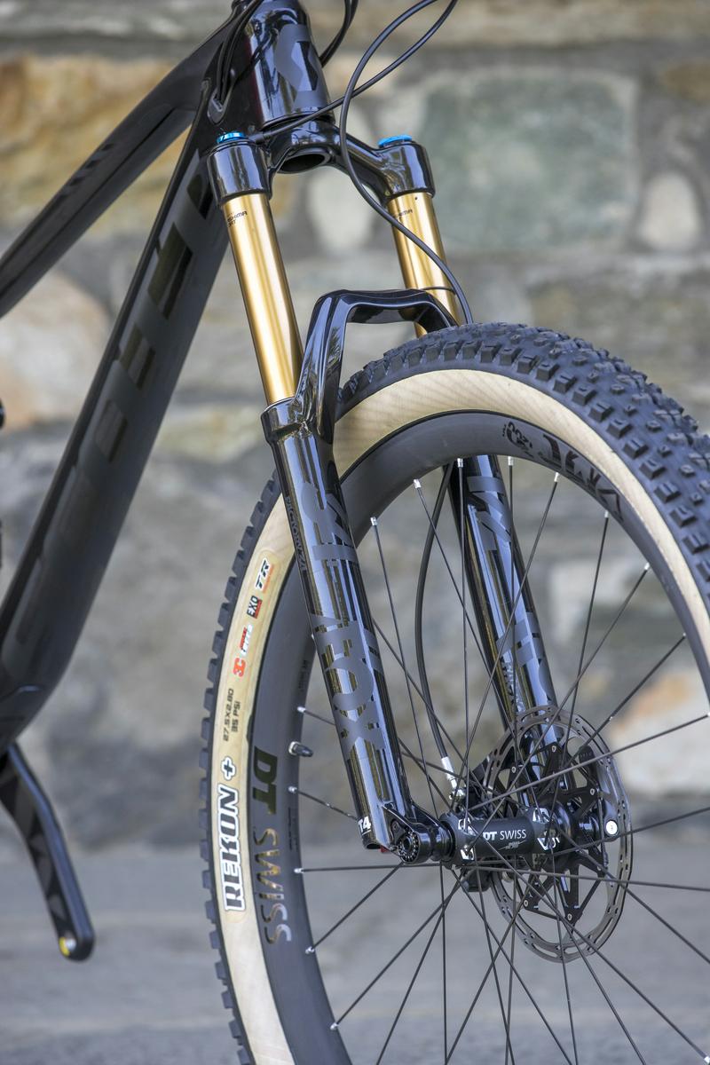 Scott Genius: Close-up view of a mountain bike front suspension fork with gold stanchions and black components, mounted on a black frame. The wheel features a thick, treaded tire with a light-colored sidewall, while the background shows a stone texture.