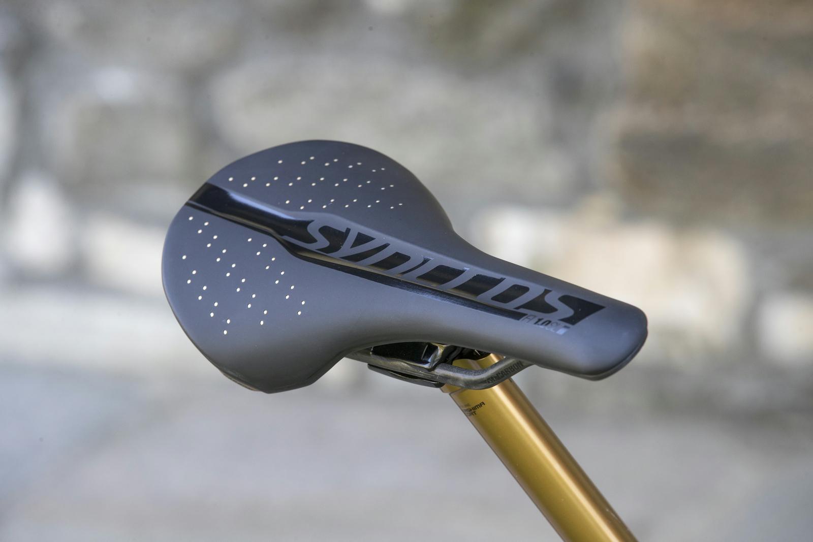 Scott Genius: A close-up view of a bicycle saddle featuring a sleek, aerodynamic design with perforations for ventilation. The saddle is primarily black with a shiny logo, and it is mounted on a gold-colored seat post against a blurred stone background.