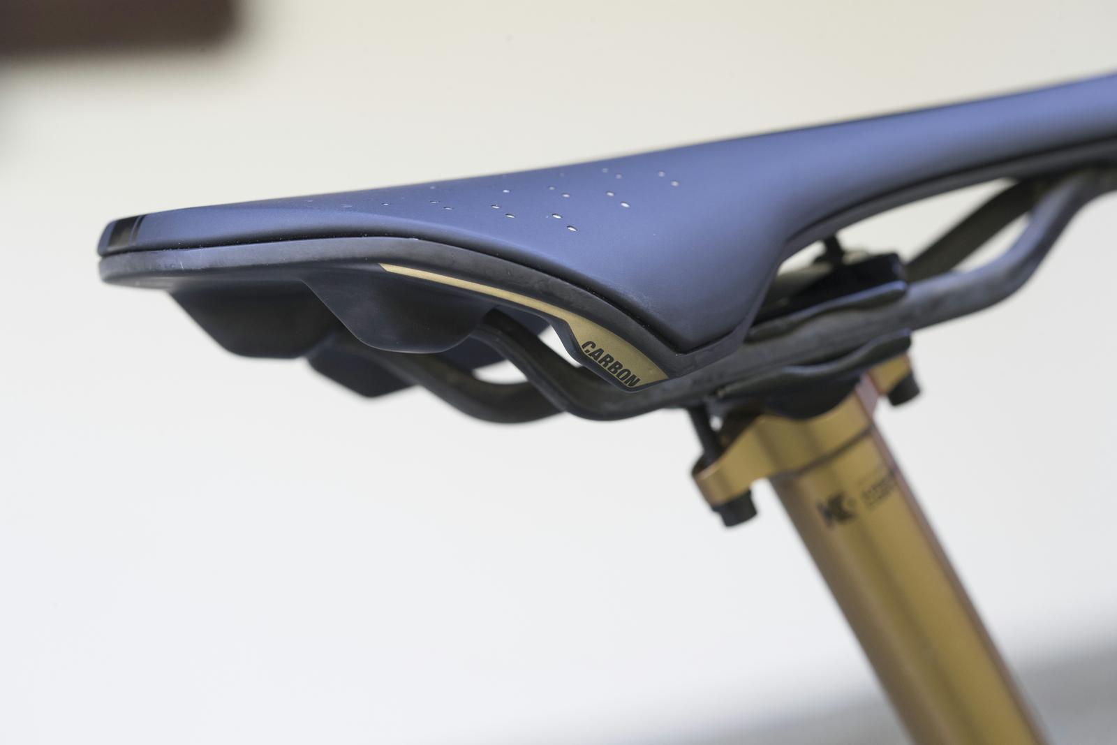Scott Genius: Close-up of a bicycle saddle, showing a sleek black design with perforations for ventilation, mounted on a gold-colored seat post. The saddle features a label indicating it is made of carbon material.