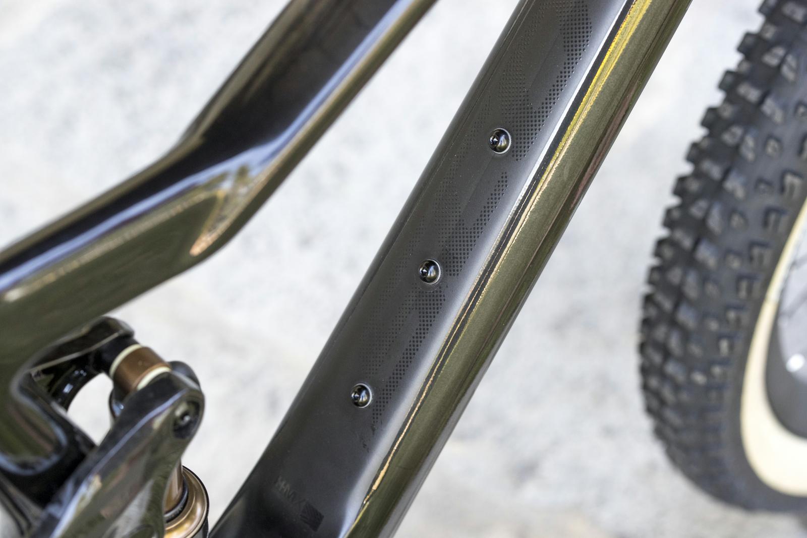 Scott Genius: Close-up image of a bicycle frame highlighting the clean design with three screws on the downtube. The frame features a sleek, dark finish with a textured pattern, showcasing the craftsmanship and attention to detail in bicycle design. A portion of the bicycle's tire is visible in the background.