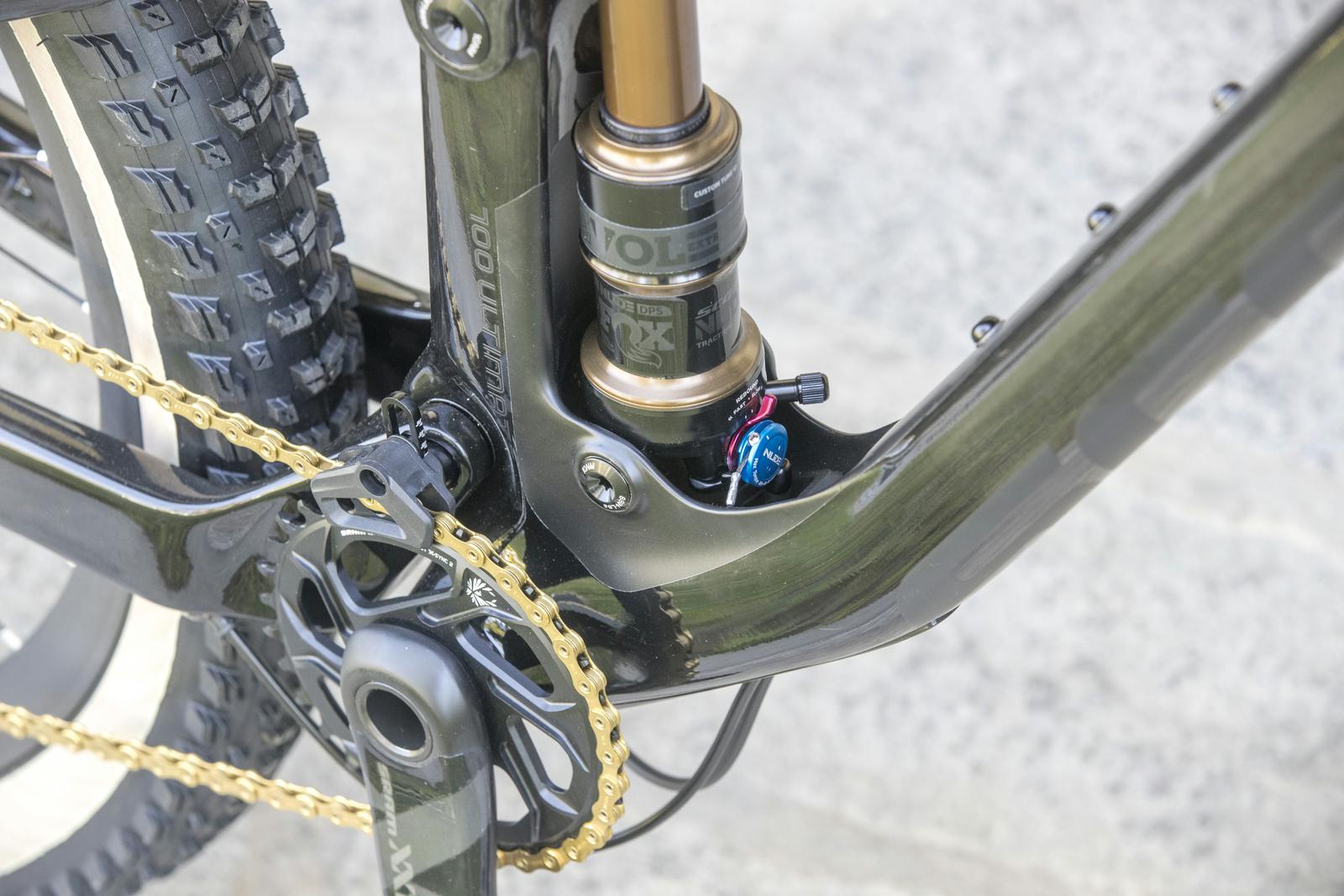 Scott Genius: Close-up view of a bicycle's drivetrain and shock system. The image showcases a black frame with a visible gold chain, a mountain bike tire, and a shock absorber with adjustment knobs, highlighting the intricate mechanical components of the bike.