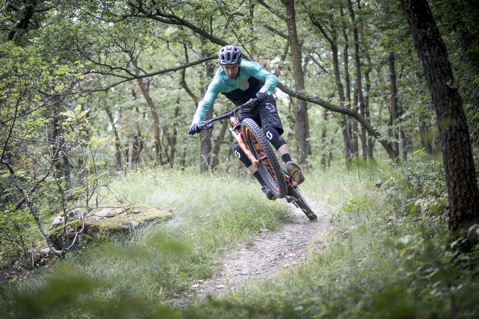 Scott Genius: A mountain biker performing a wheelie on a forest trail, surrounded by greenery and trees. The rider is wearing a helmet and protective gear, showcasing dynamic movement and skill on the bike.