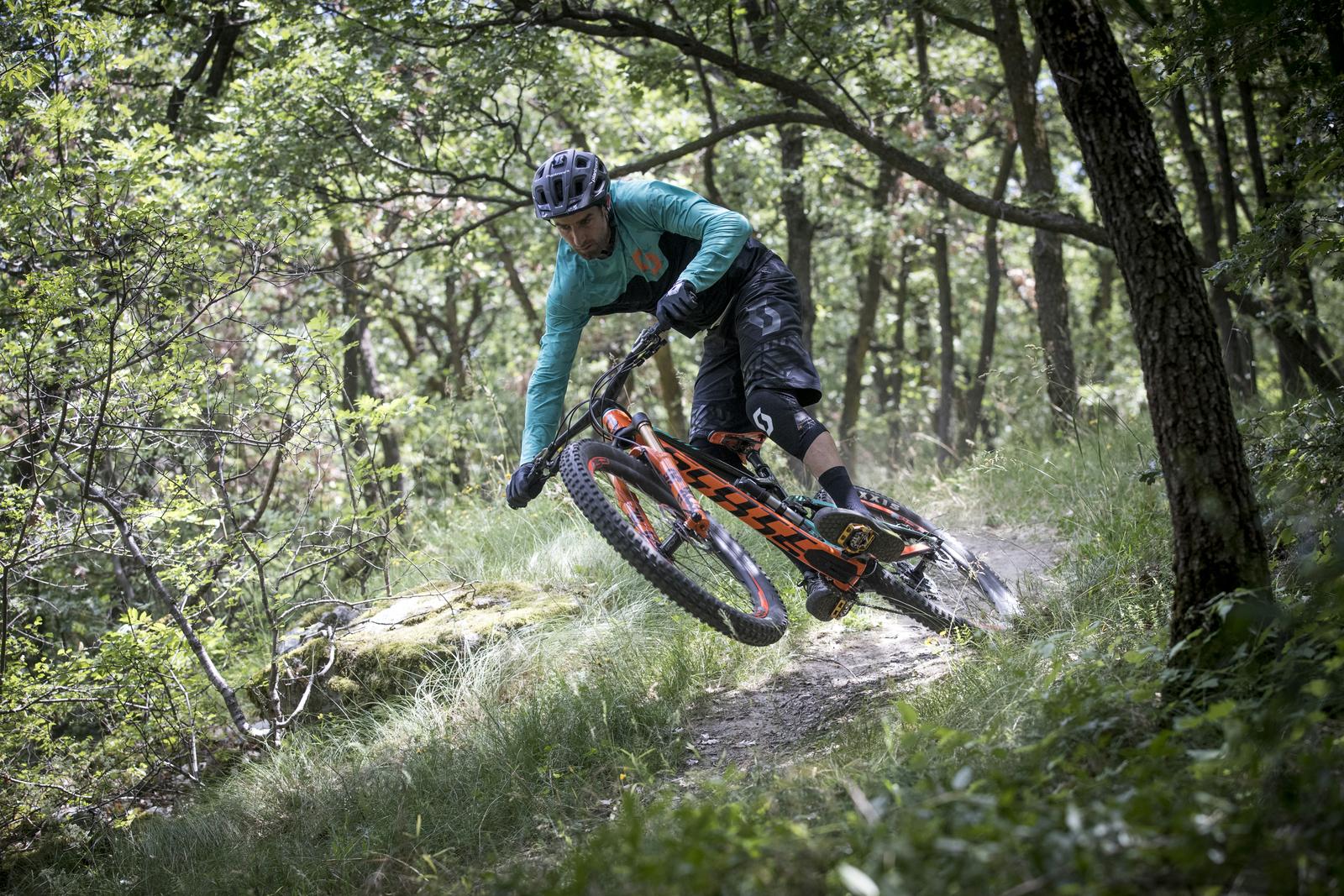 Scott Genius: A mountain biker performing a jump on a dirt trail surrounded by lush green trees and underbrush. The rider is dressed in a long-sleeve turquoise shirt and black shorts, focused on maneuvering the bike mid-air.