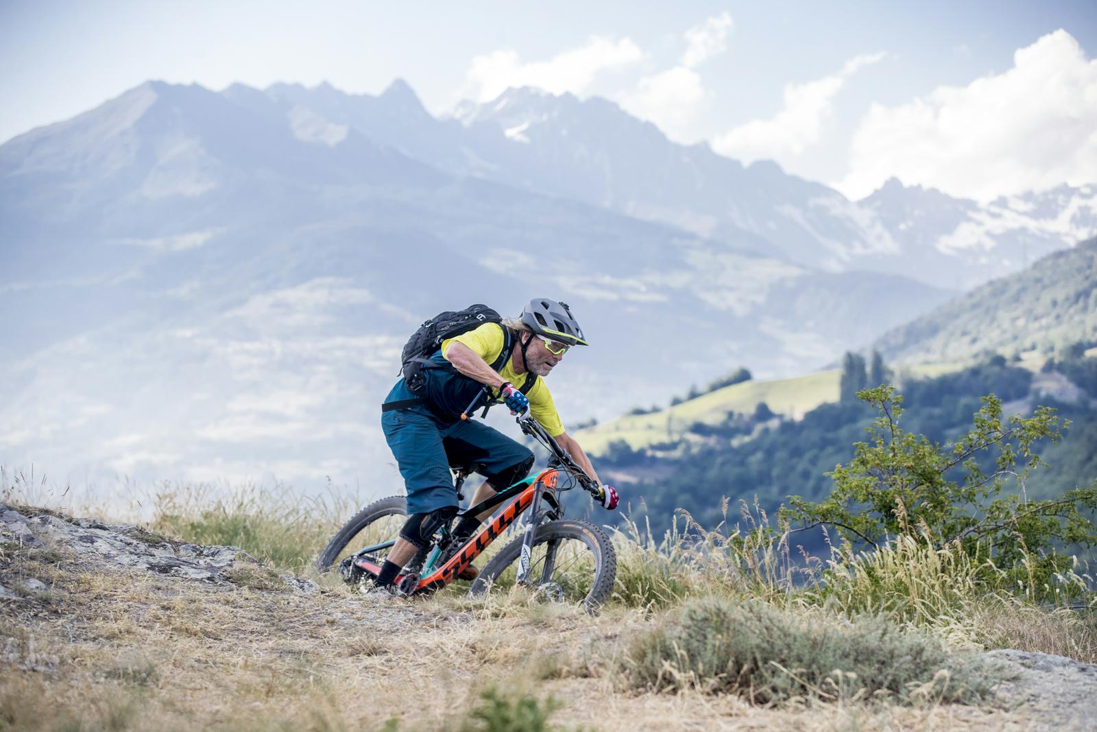 Scott Genius: A mountain biker in a bright yellow shirt and blue shorts navigates a rocky trail surrounded by mountains and greenery. He is leaning forward on his bike, wearing a helmet and sunglasses, with a backpack secured on his back. The landscape features hills and distant snow-capped peaks under a clear sky.