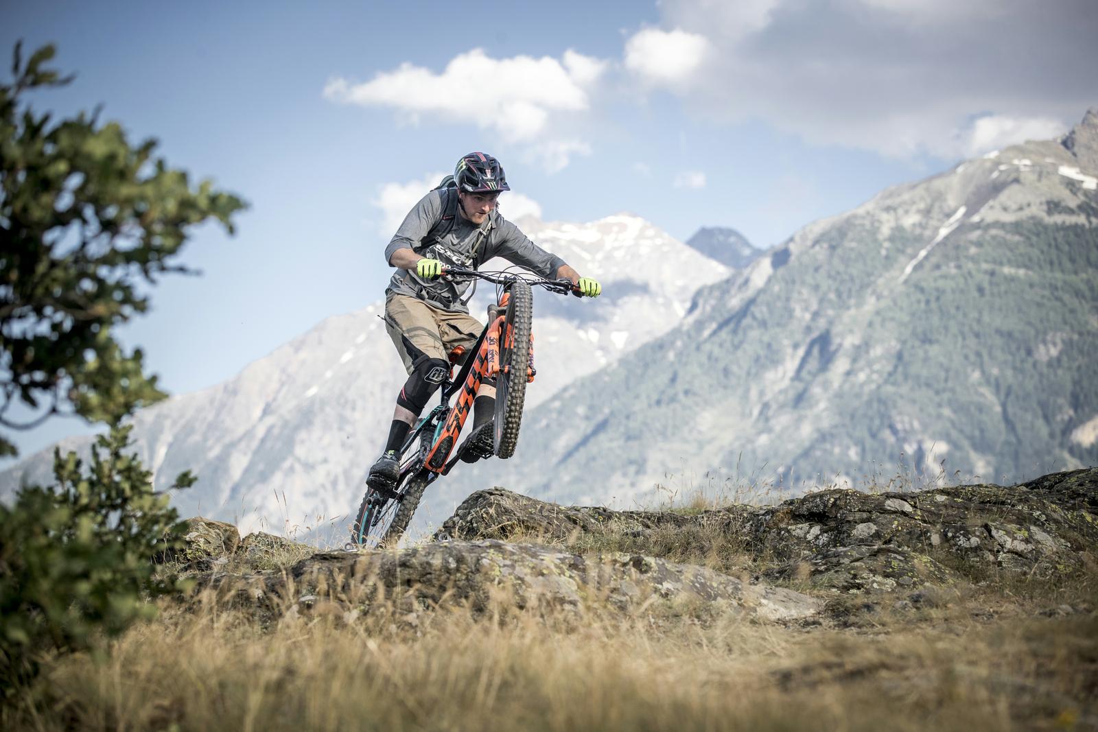 Scott Genius: A mountain biker performing a wheelie on rocky terrain, surrounded by grass and mountains under a partly cloudy sky. The cyclist is wearing a helmet, gloves, and protective gear while demonstrating skill on a trail.