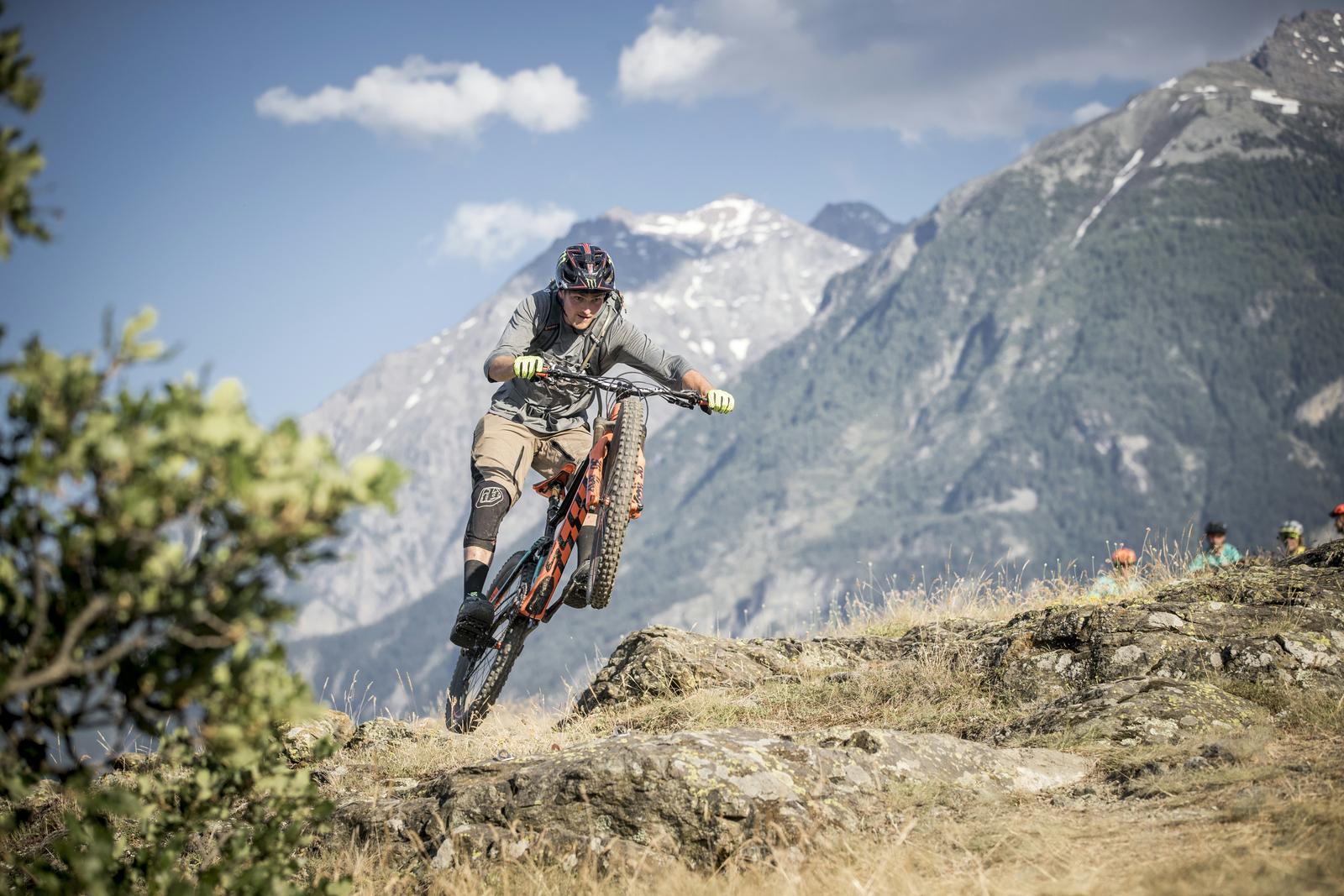 Scott Genius: A mountain biker performing a trick on rocky terrain, with a backdrop of mountains and a clear blue sky. The biker is dressed in a gray shirt and shorts, wearing a helmet and gloves, and is focused on maintaining balance while navigating the challenging landscape. The scene captures the adventurous spirit of mountain biking in a natural setting.