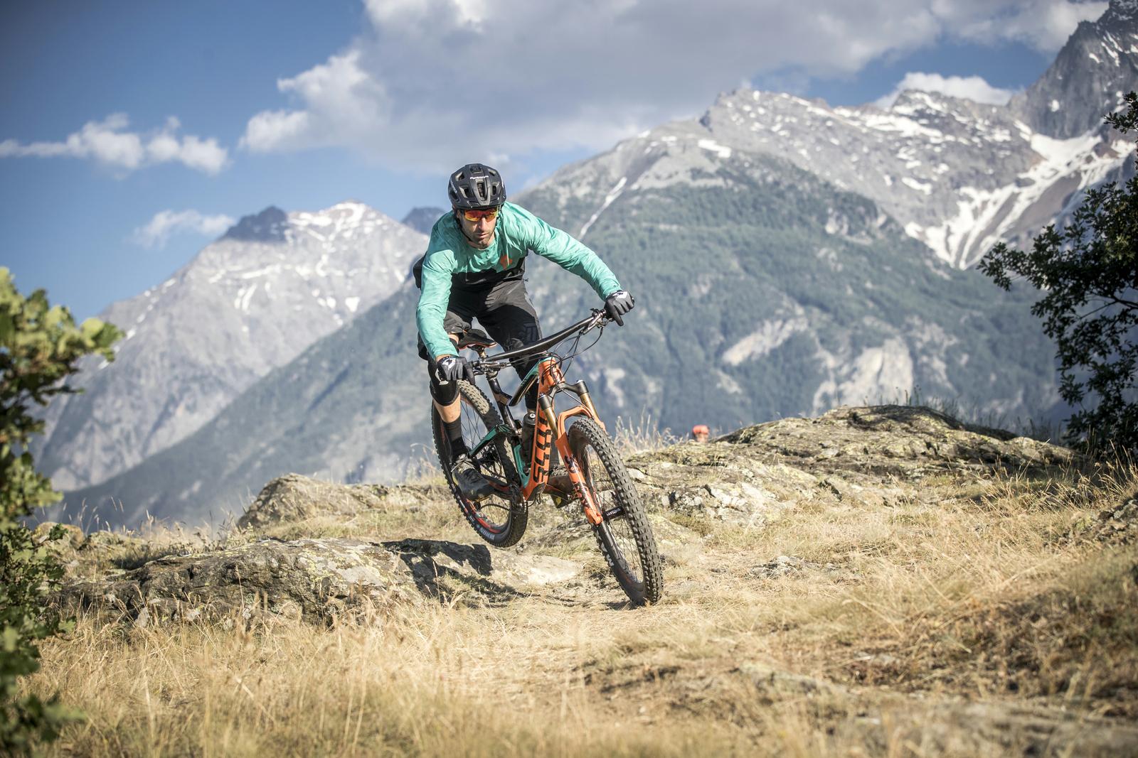 Scott Genius: A person in a turquoise jacket and black shorts riding a mountain bike over rocky terrain with a panoramic view of mountains in the background under a blue sky with scattered clouds.