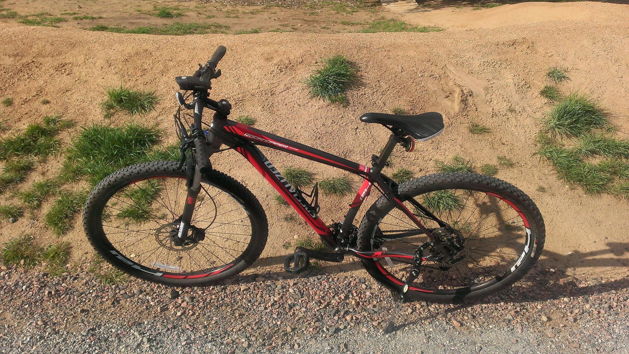 Specialized Rockhopper Comp 29: A red and black mountain bike is leaning on a gravel path, surrounded by patches of grass and dirt. The bike features thick tires, a black seat, and visible gear components.