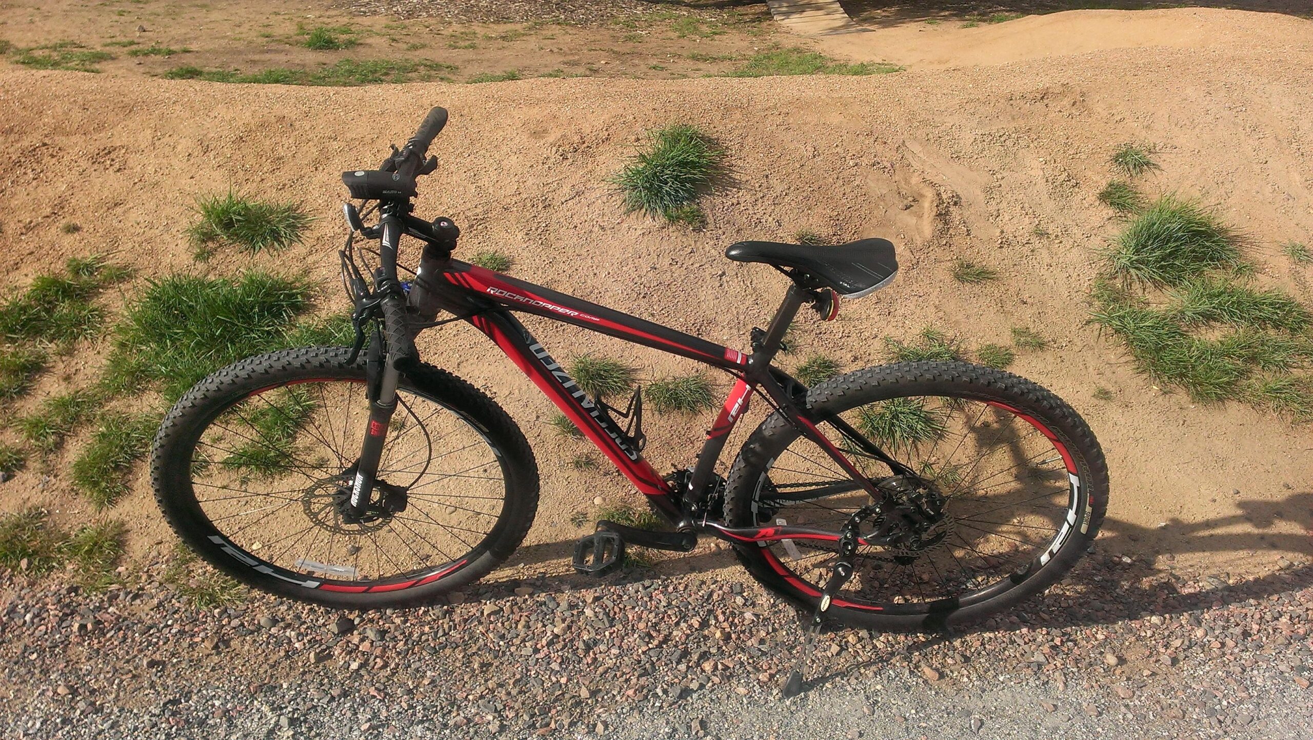 Specialized Rockhopper Comp 29: A red and black mountain bike resting on a gravel path, with grassy areas and dirt mounds in the background. The bike features wide tires, a sturdy frame, and visible components like the handlebars and pedals.