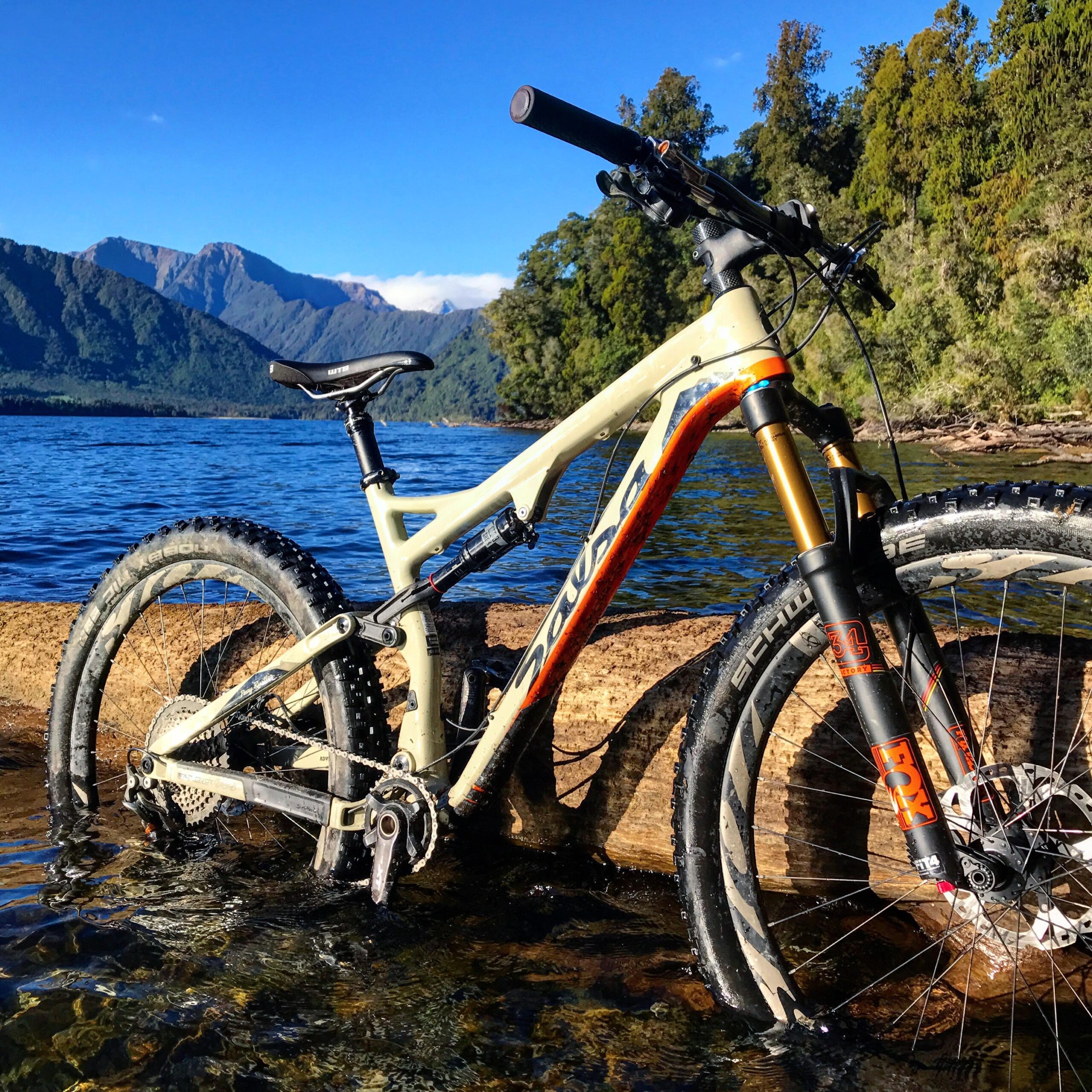 Salsa Pony Rustler Carbon XO1: A mountain bike partially submerged in clear water at the edge of a lake, surrounded by lush greenery and mountains in the background under a bright blue sky.