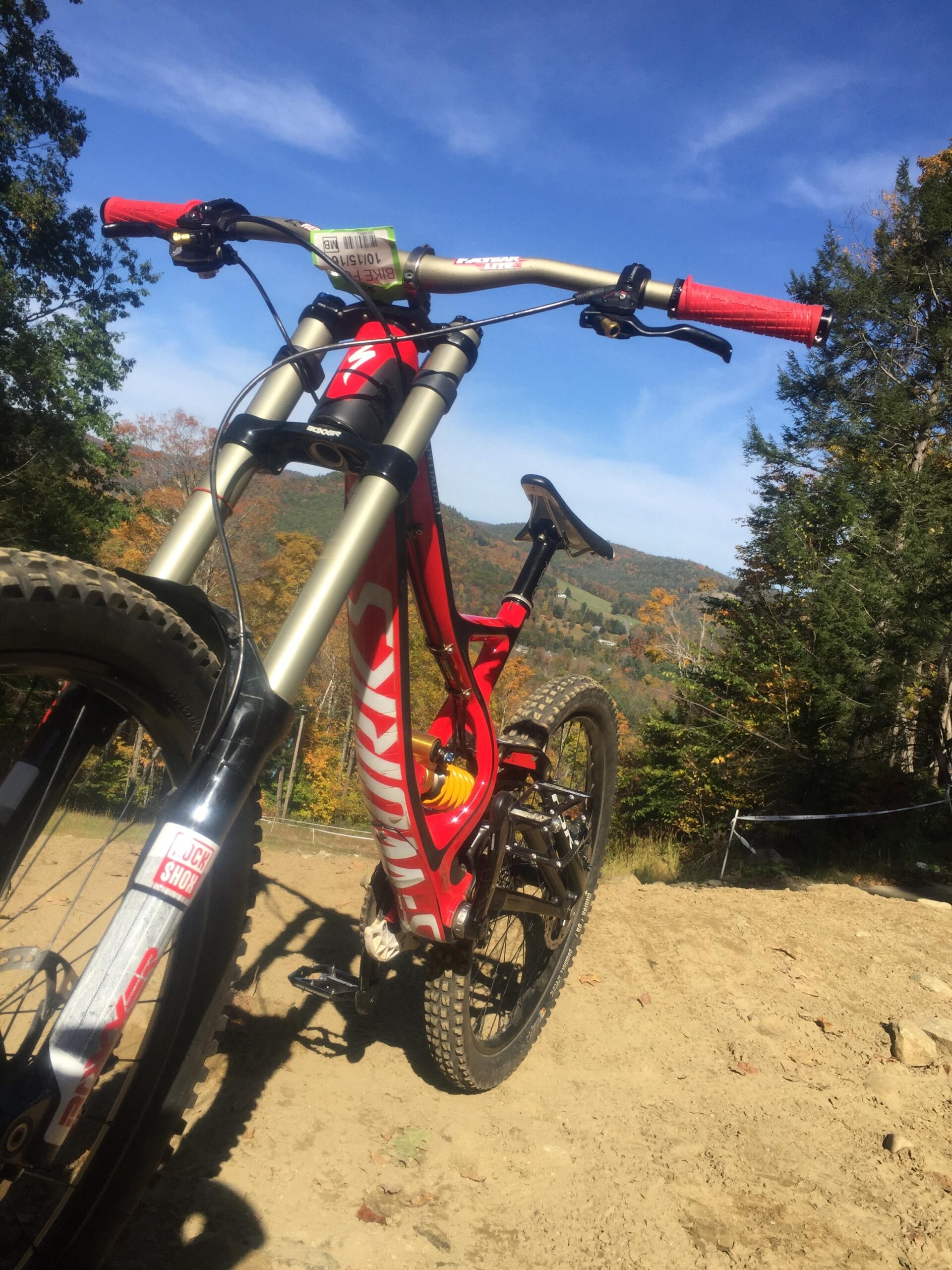 Specialized S-Works Demo 8 Carbon: Alt text: A close-up view of a mountain bike positioned on a dirt trail, with vibrant autumn foliage in the background. The bike features a red frame and handle grips, with a visible suspension system and thick tires suitable for off-road riding. The sky is clear with scattered clouds.