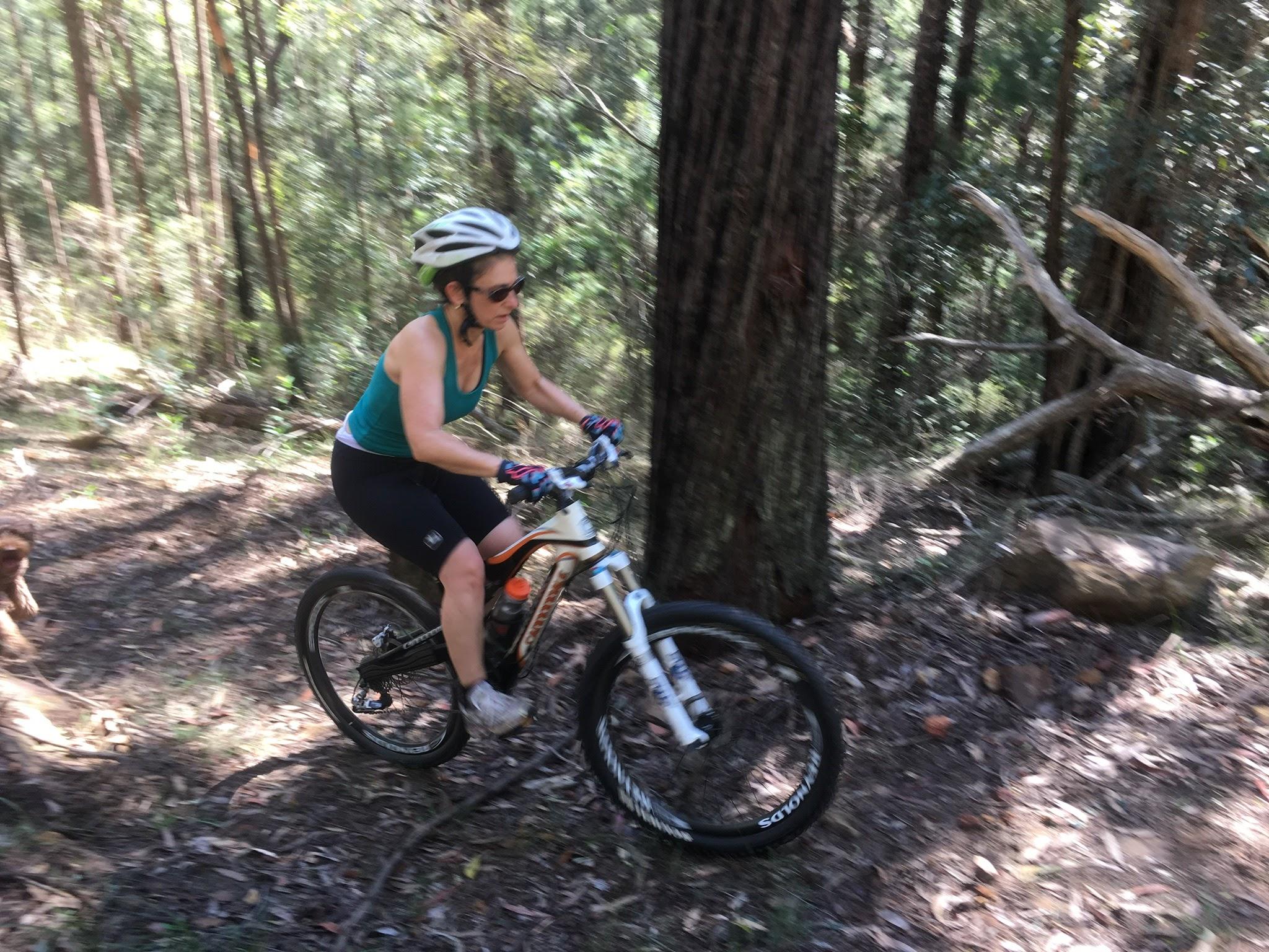 A woman riding a mountain bike along a forest trail, surrounded by tall trees and foliage. She is wearing a helmet and sunglasses, dressed in a tank top and cycling shorts. The scene captures a sense of motion as she navigates through the natural landscape. Trailshare Kulnura mountain bike trail.