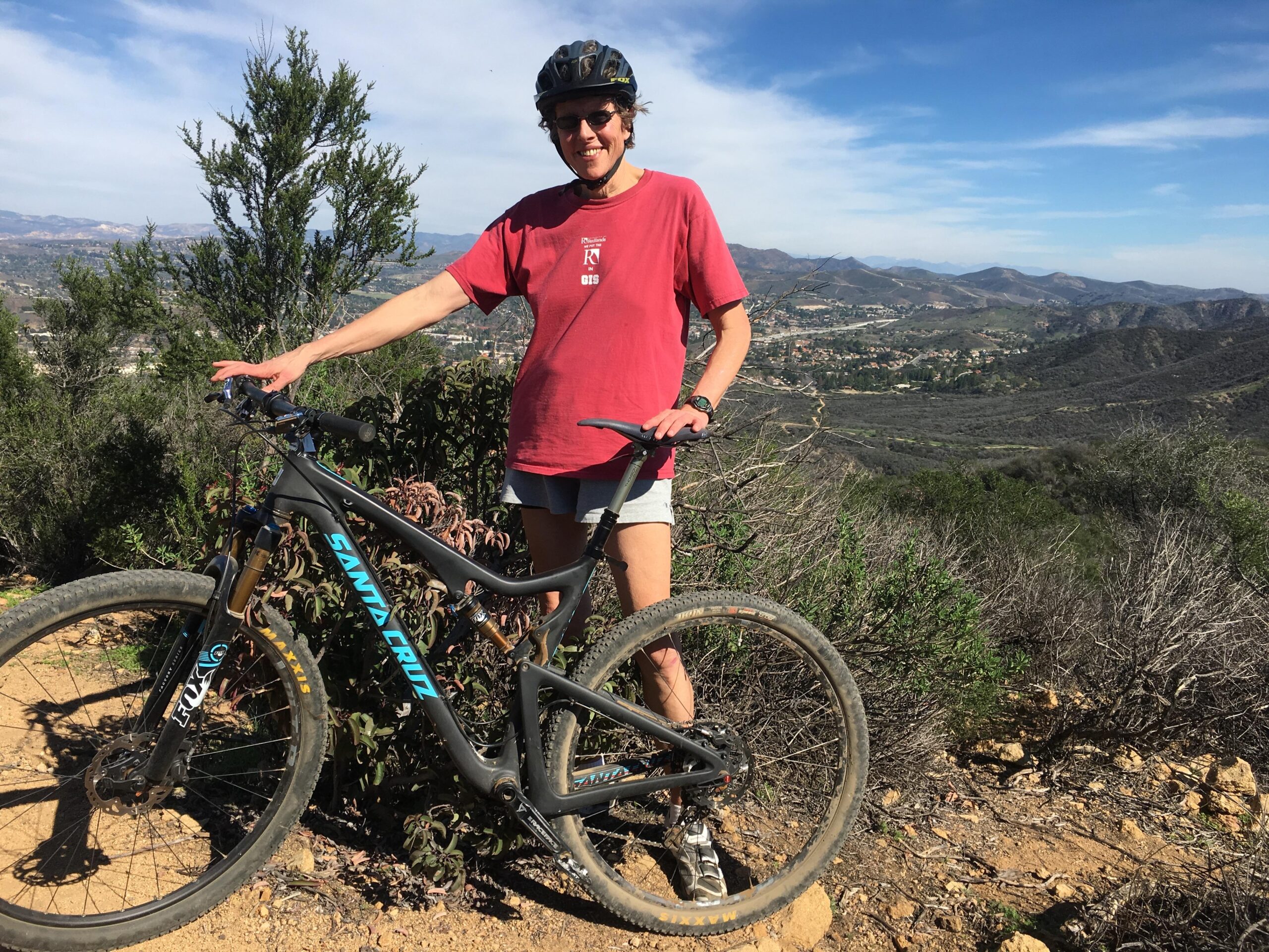 Santa Cruz Tallboy Carbon: A person wearing a helmet and a red t-shirt stands next to a mountain bike on a dirt trail with a scenic view of mountains and valleys in the background. The landscape features some greenery and distant hills under a partly cloudy sky.