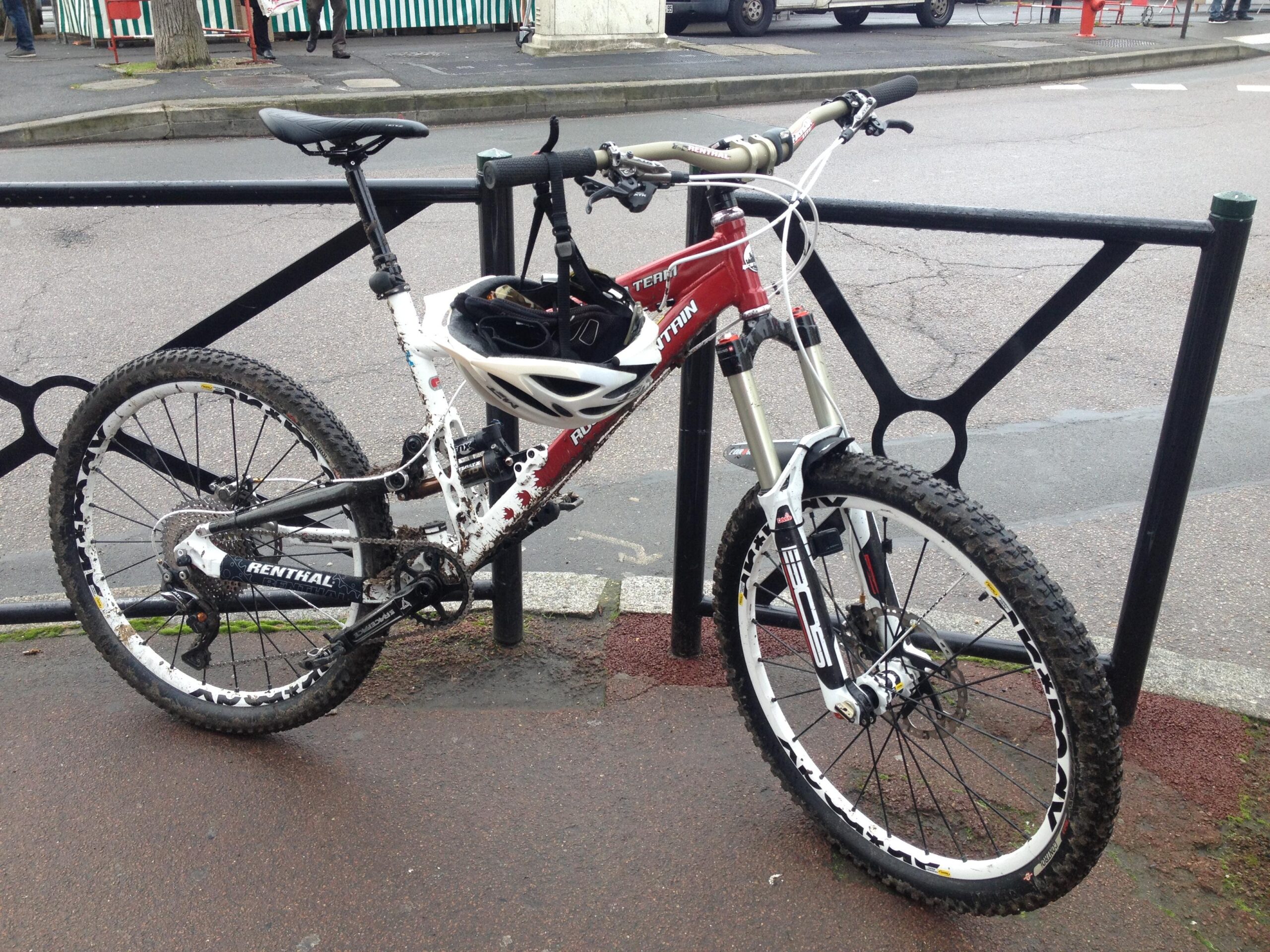 Rocky Mountain Slayer: A red and white mountain bike with distinct branding, resting against a black bike rack. The bike shows signs of use, with dirt on the tires and frame, and a helmet secured on the handlebars. In the background, a partially visible street scene includes buildings and pedestrians.