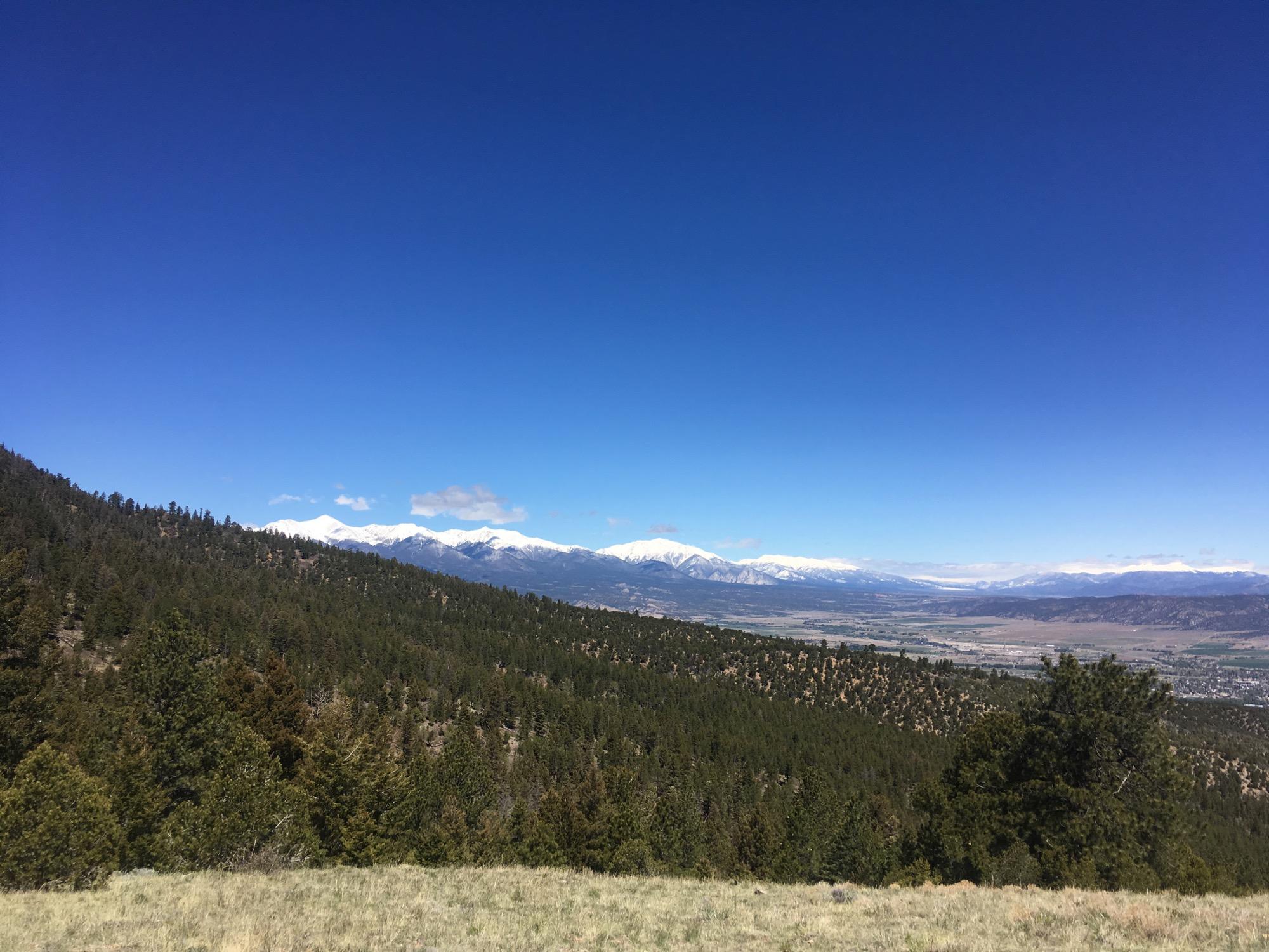 A panoramic view of a mountainous landscape featuring snow-capped peaks in the distance, lush green forests in the foreground, and a clear blue sky with a few scattered clouds. Rainbow Trail: Methodist Mountain Thd to Bear Creek Thd mountain bike trail.