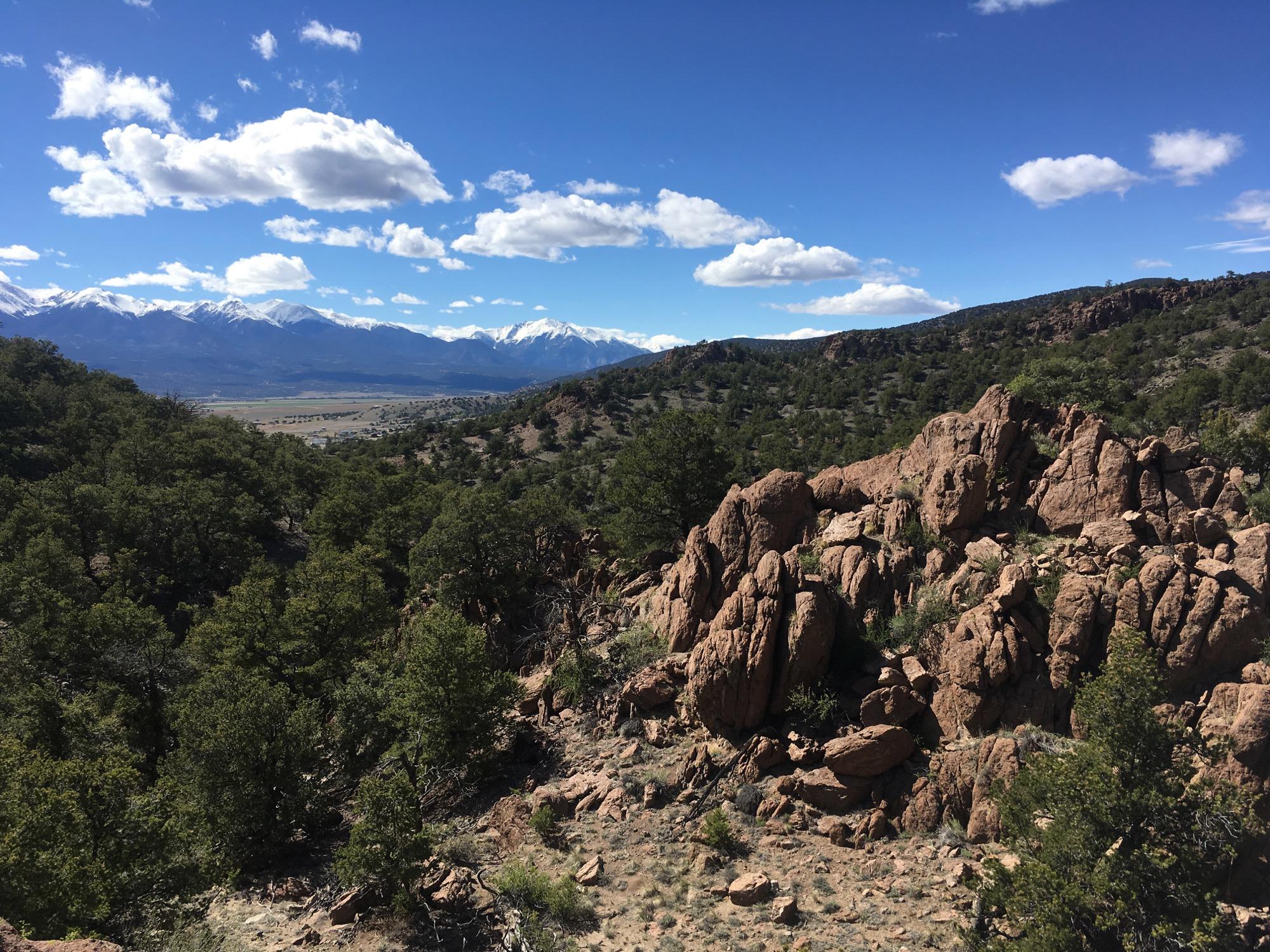 A panoramic view of a mountainous landscape featuring rocky formations in the foreground, lush green trees in the midground, and snow-capped peaks in the distance under a partly cloudy blue sky. North Backbone mountain bike trail.